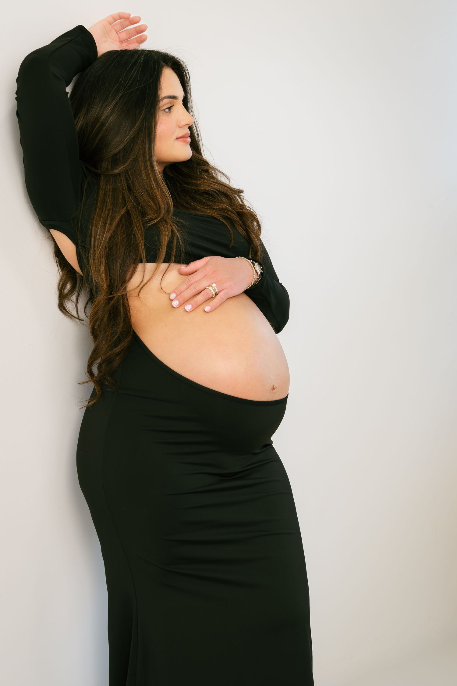 Pregnant woman in black dress cradling baby bump, posing against white wall with long dark hair.