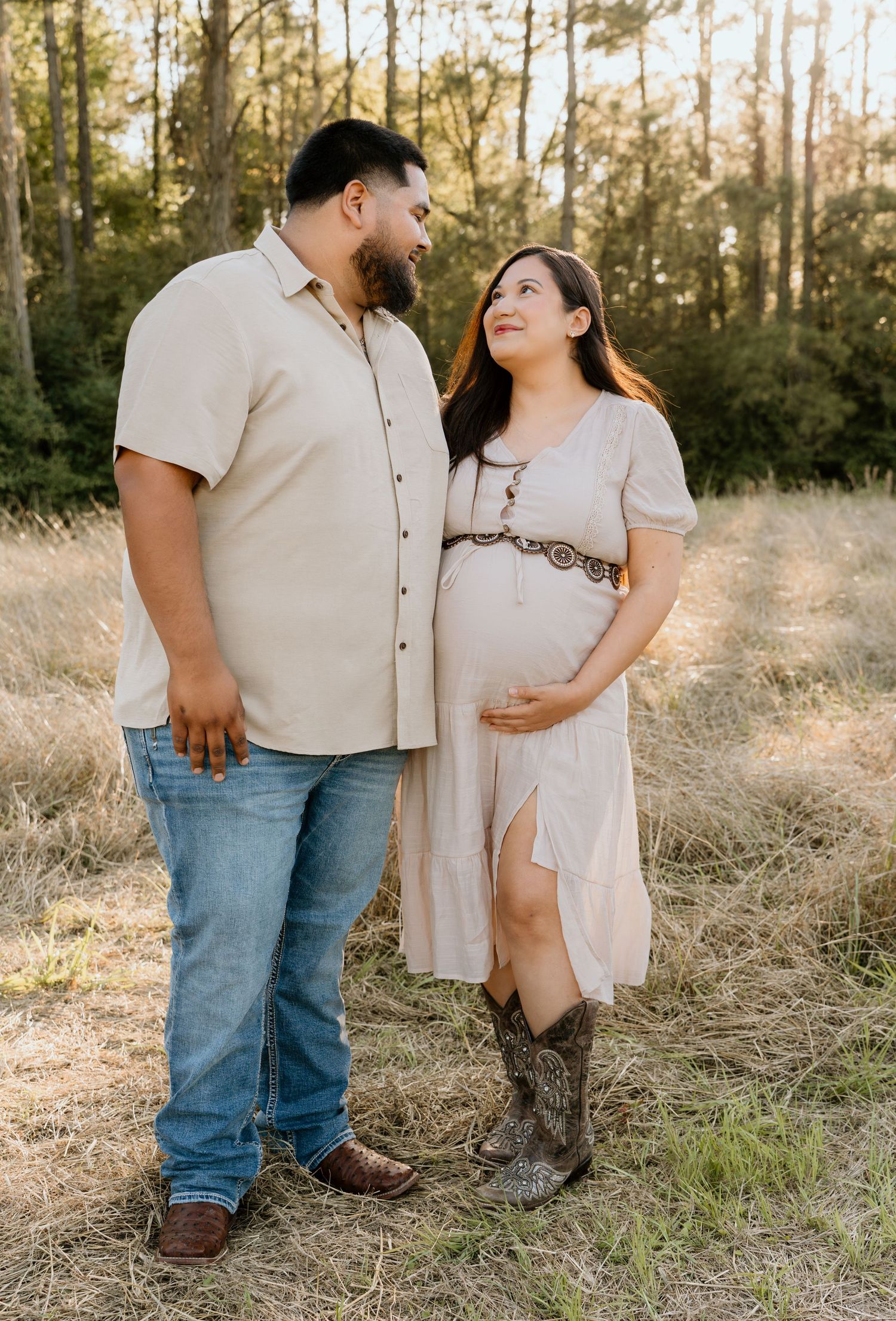 Couple poses in a field, pregnant woman in beige dress and cowboy boots, man in khaki shirt and jeans, surrounded by trees.