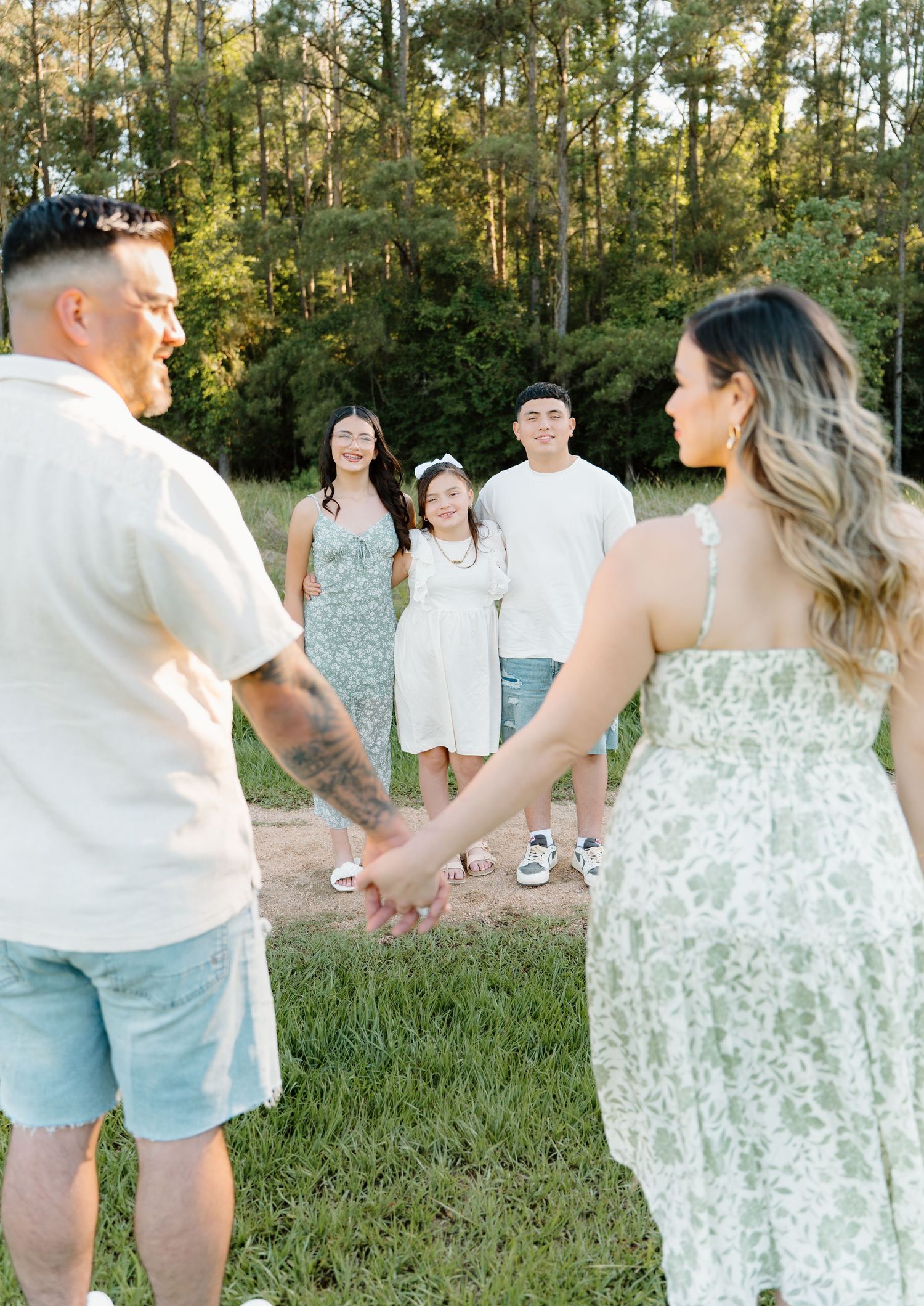 Family holding hands outdoors with three children smiling in background, surrounded by lush green trees.