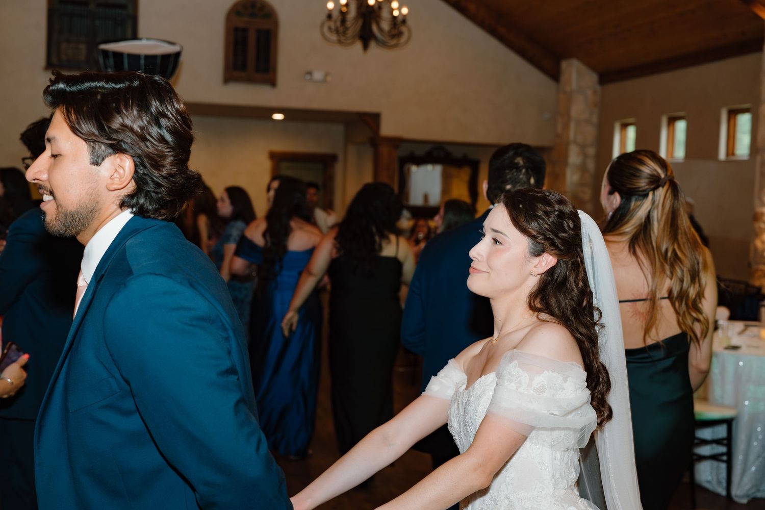 Bride in off-shoulder wedding gown dancing with groom in navy suit at elegant indoor wedding reception.