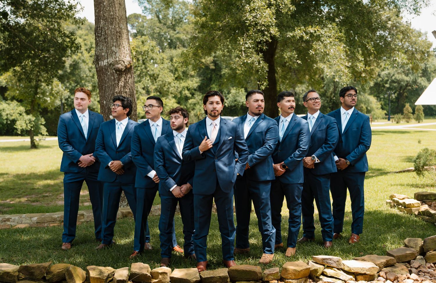Groomsmen in navy blue suits posing outdoors on a green lawn with stone wall and trees at a wedding.