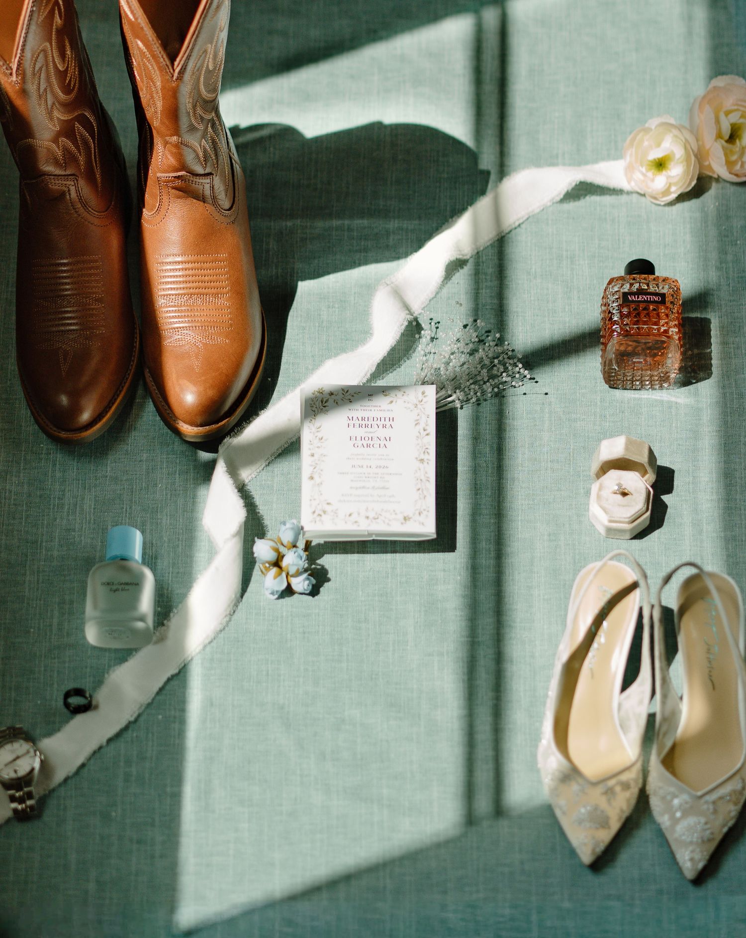 Flat lay of wedding details including cowboy boots, heels, perfume, watch, florals, and invitation on green fabric.