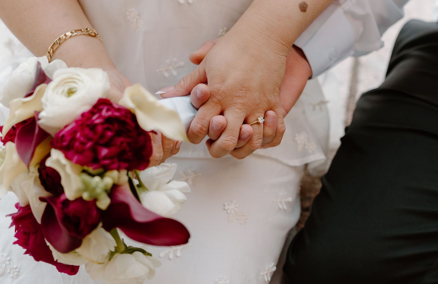 Close-up of newlyweds holding hands showing wedding rings with red peony and white floral bouquet.