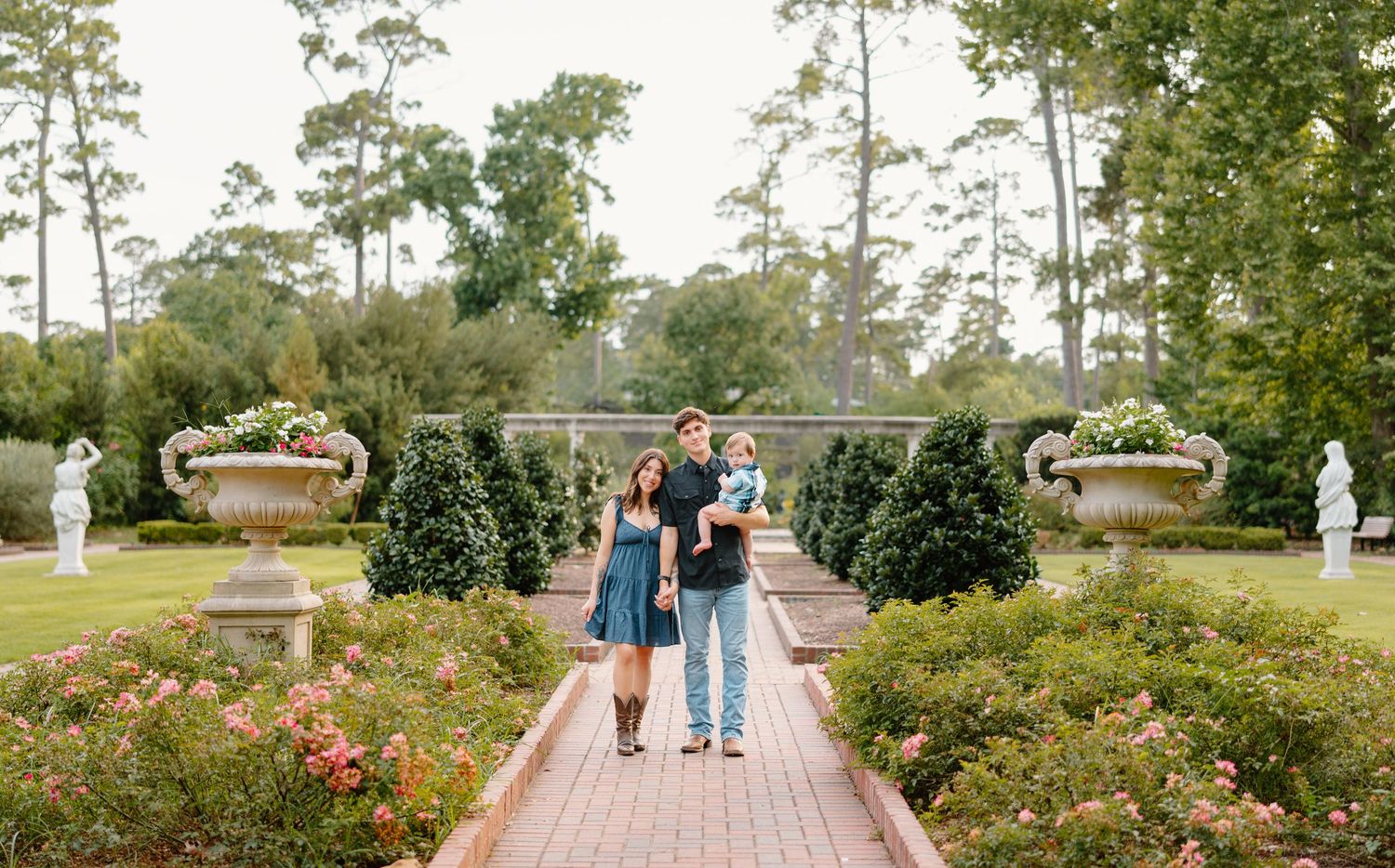Family of three posing in a lush formal garden with rose bushes, stone urns, and tall trees in the background.