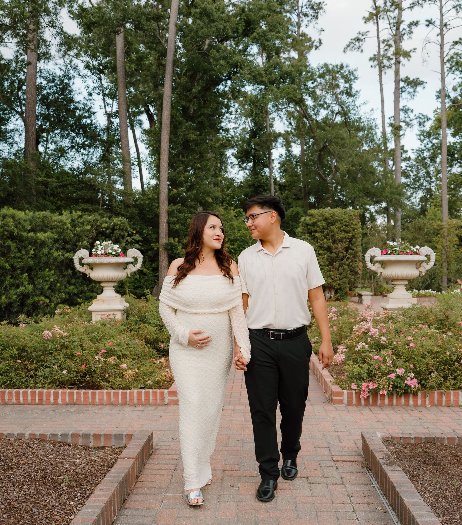 Pregnant couple walking hand in hand through a garden, woman in white dress, man in white polo and black pants.