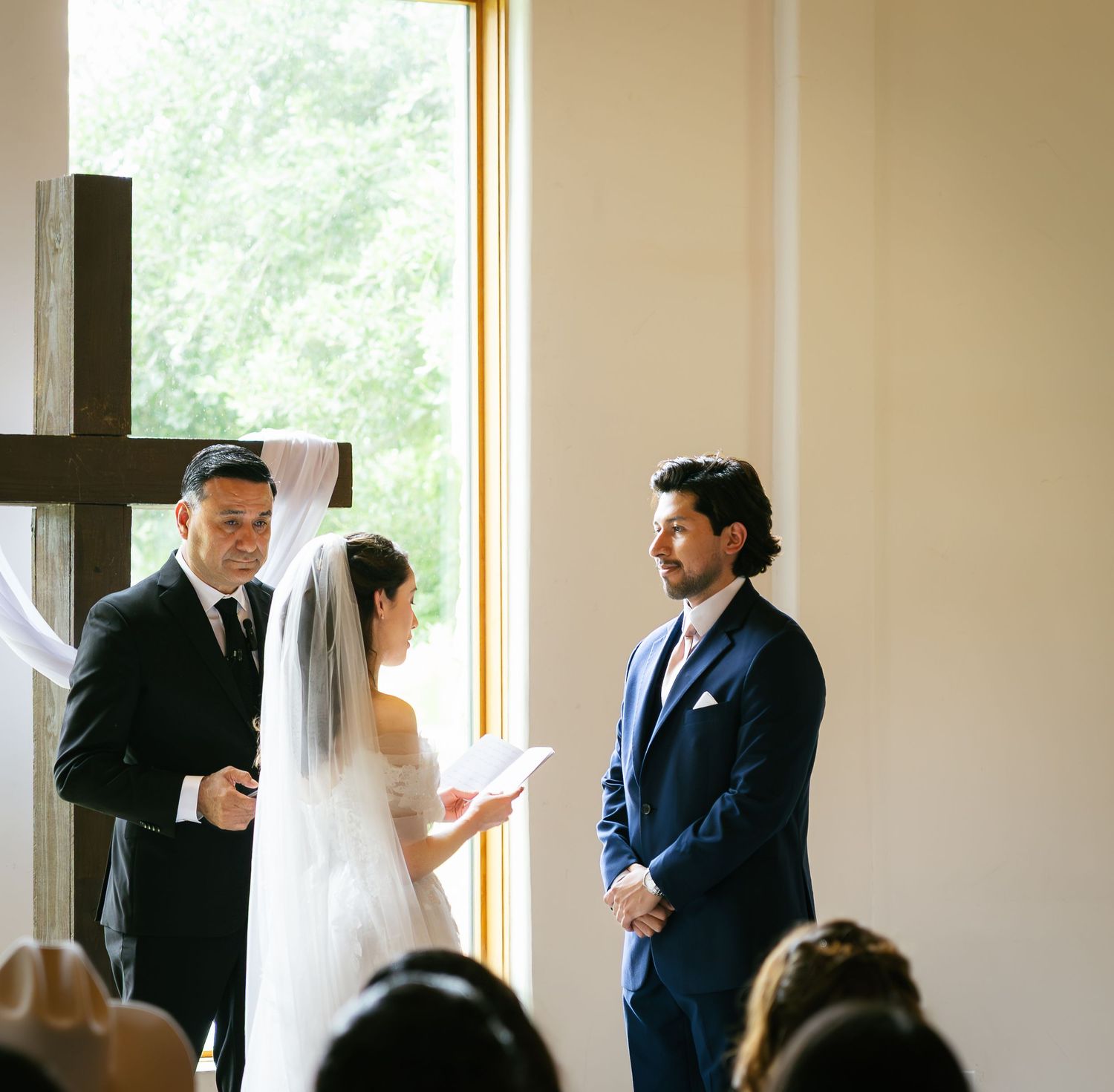 Bride and groom exchange vows at elegant wedding ceremony with wooden cross and large window backdrop.