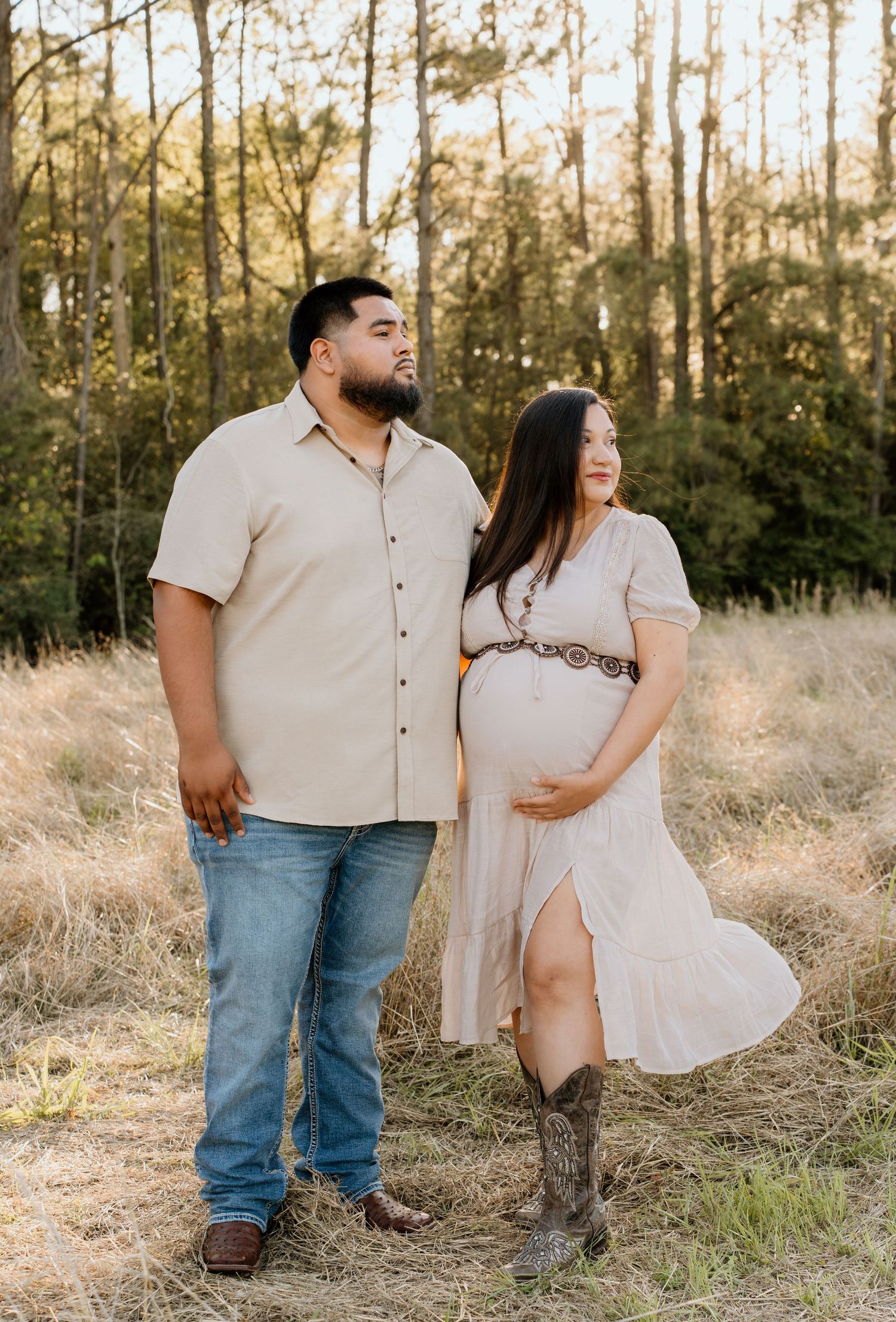 Expecting couple in matching beige outfits posing in golden field at sunset.