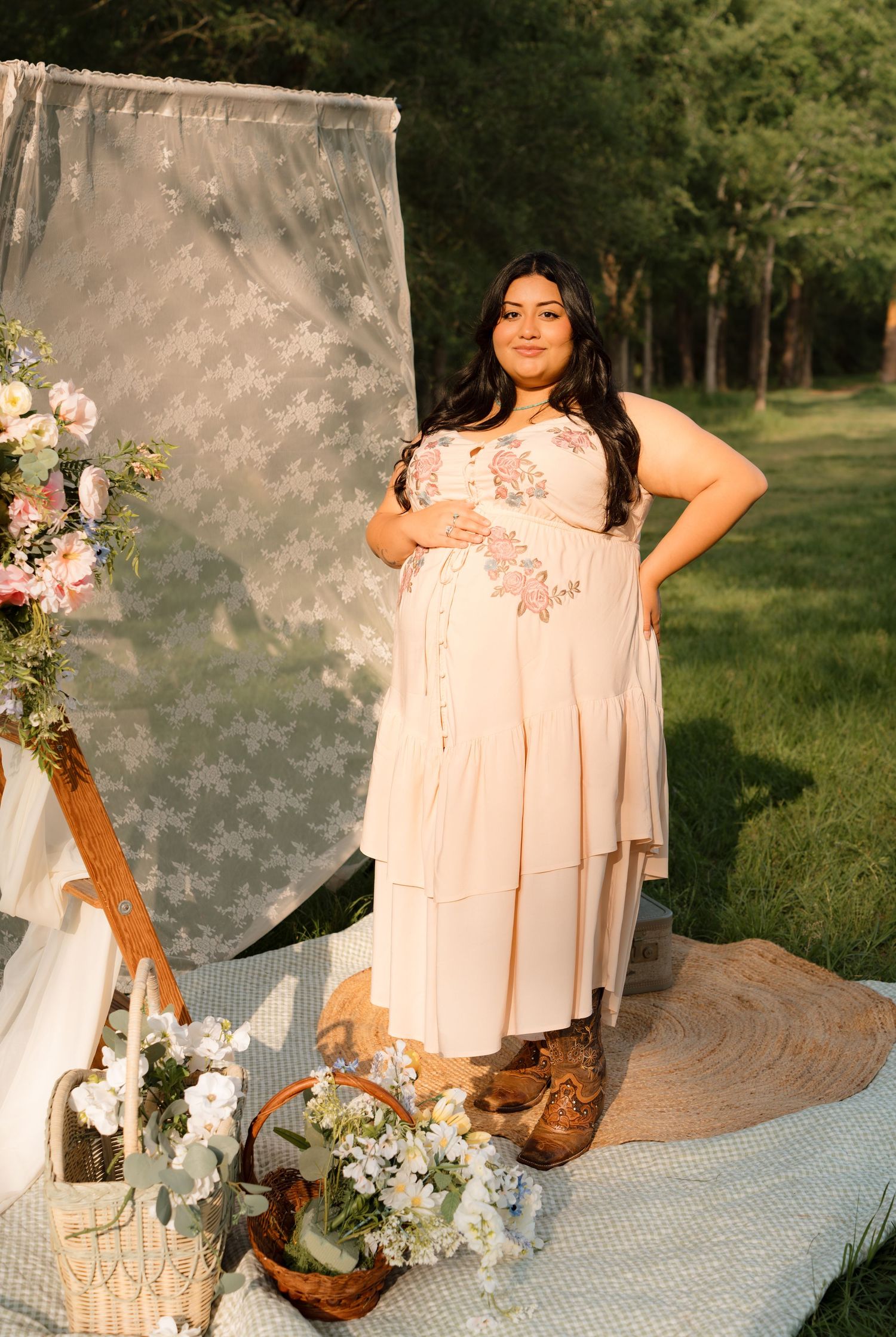 Pregnant woman in blush floral dress posing outdoors at a boho maternity photoshoot with flowers and greenery.