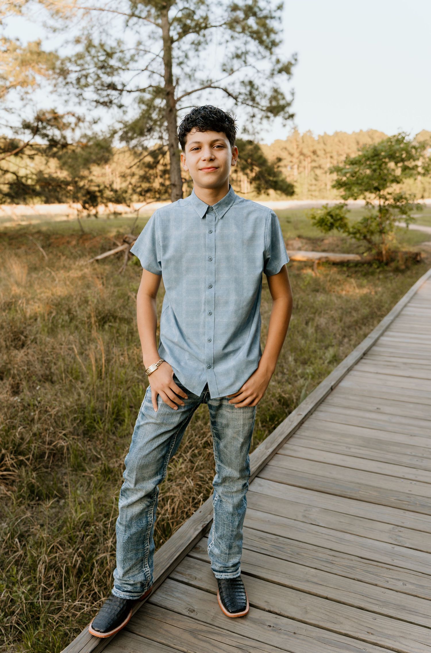 Teen boy in blue shirt and jeans stands on wooden boardwalk surrounded by trees and meadow in golden hour light.