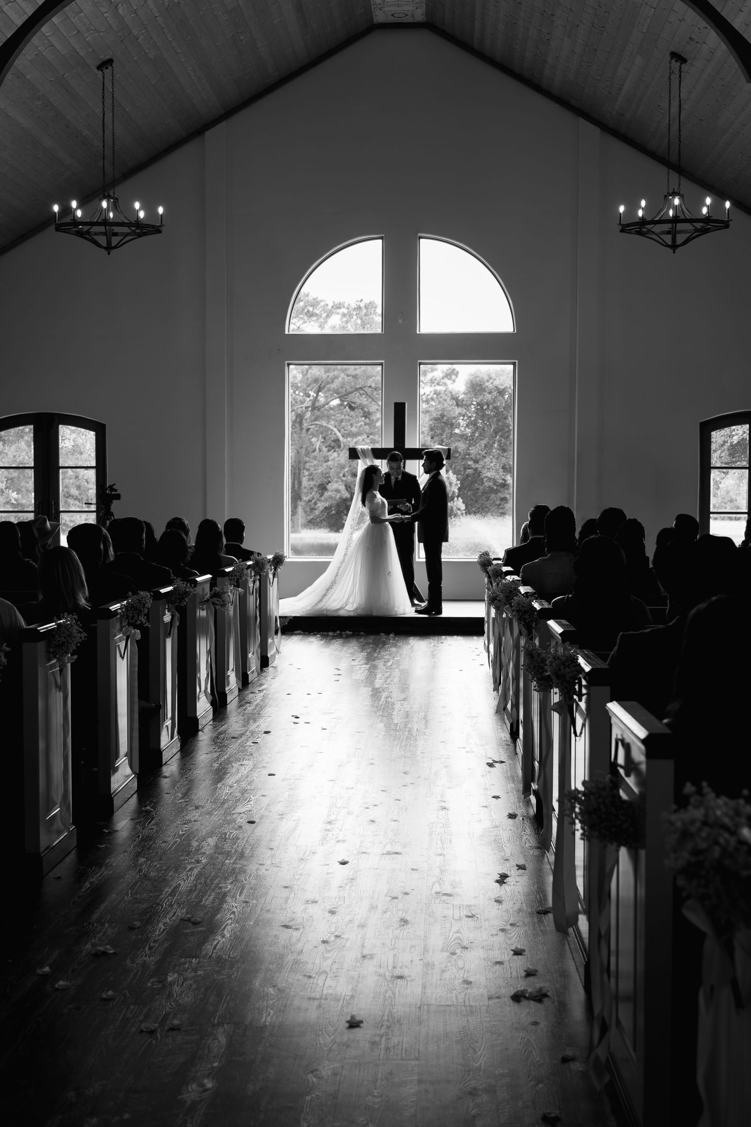 Black and white photo of bride and groom at altar in elegant chapel with wooden pews and arched window.