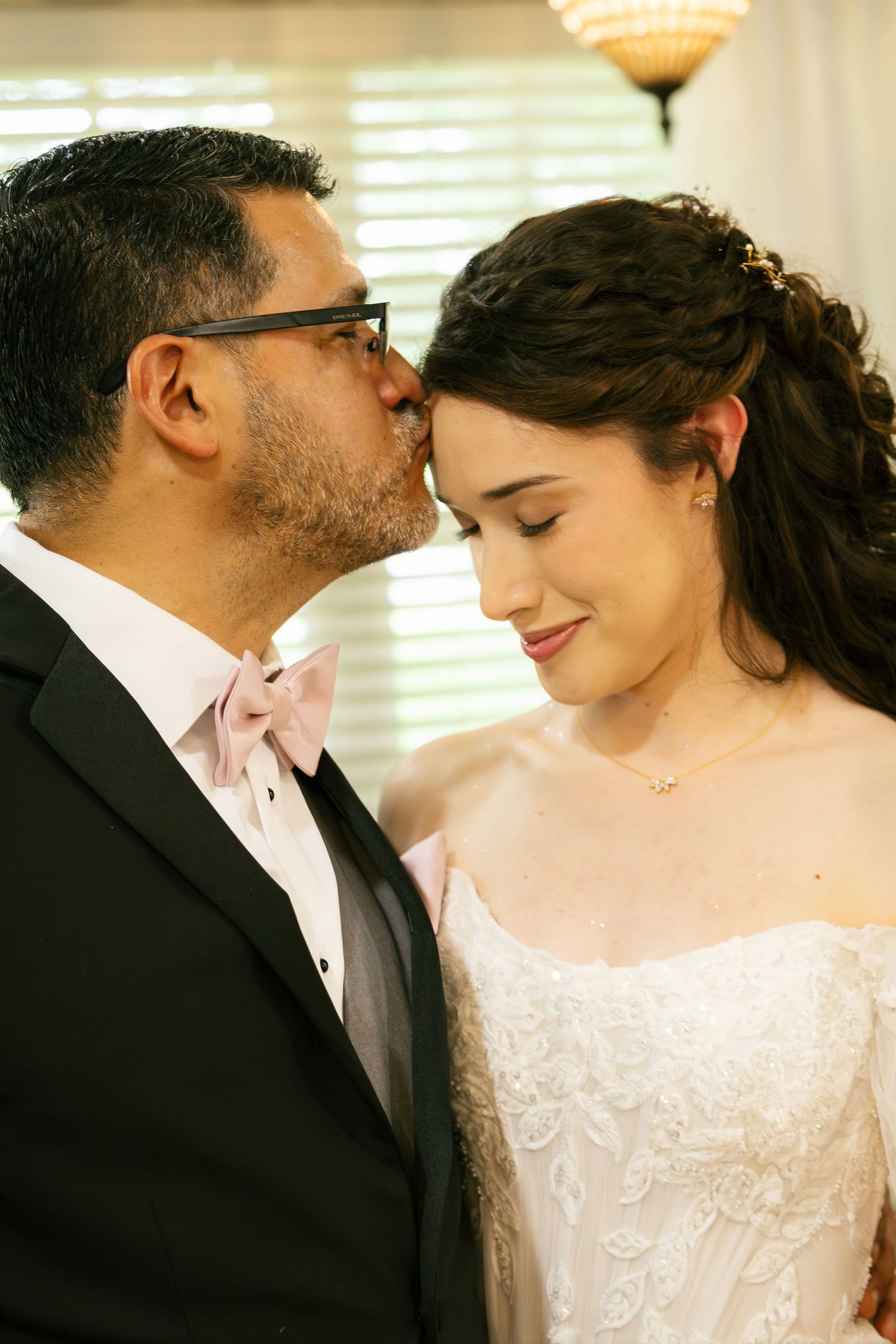 Father of bride in black tuxedo with pink bow tie kissing bride on forehead during wedding day.