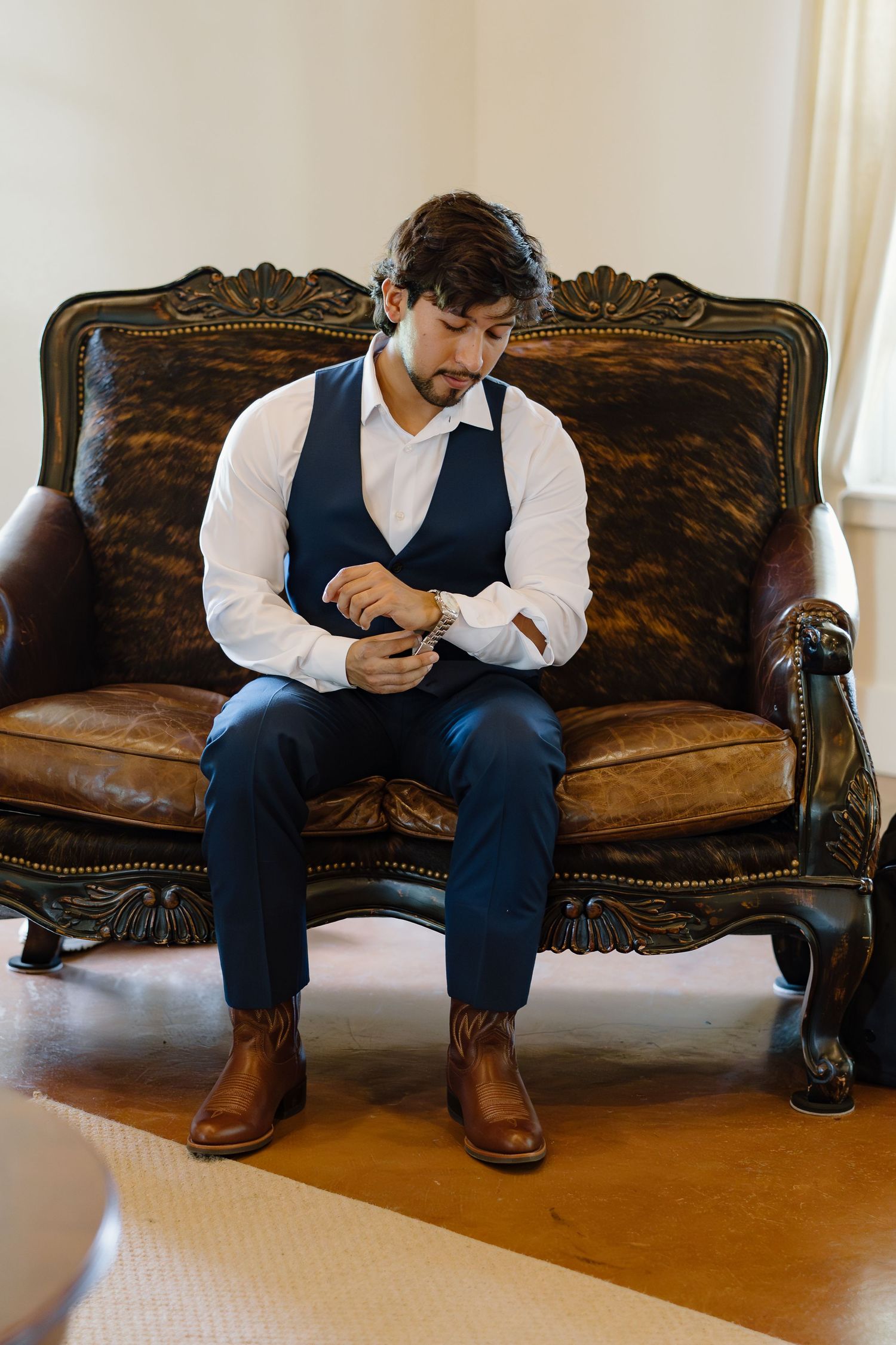 Groom in navy vest and white shirt adjusting cufflinks while seated on ornate leather sofa before wedding.