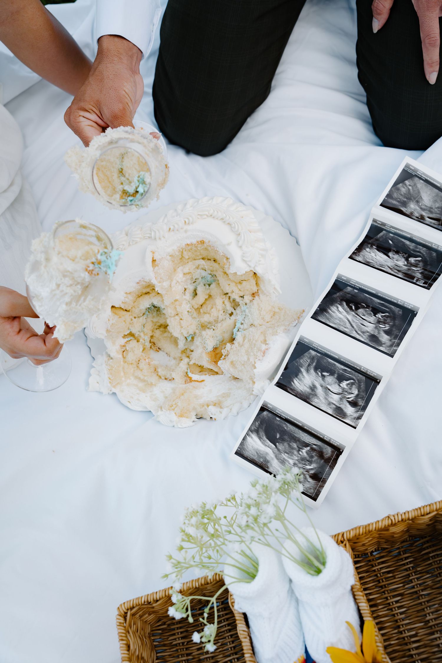 Couple holding glasses near ultrasound photos, baby socks, and a decorated cake on white fabric at a gender reveal.