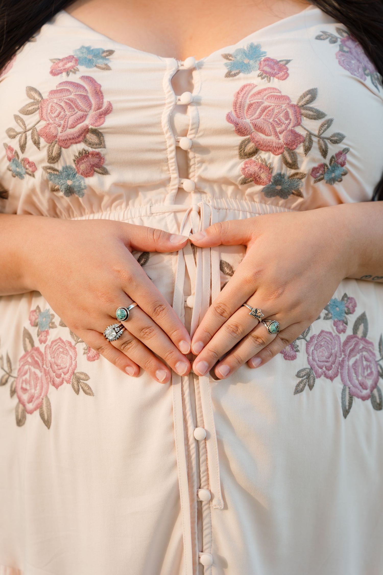 Pregnant woman in floral embroidered white dress forming heart shape on baby bump with ringed hands.