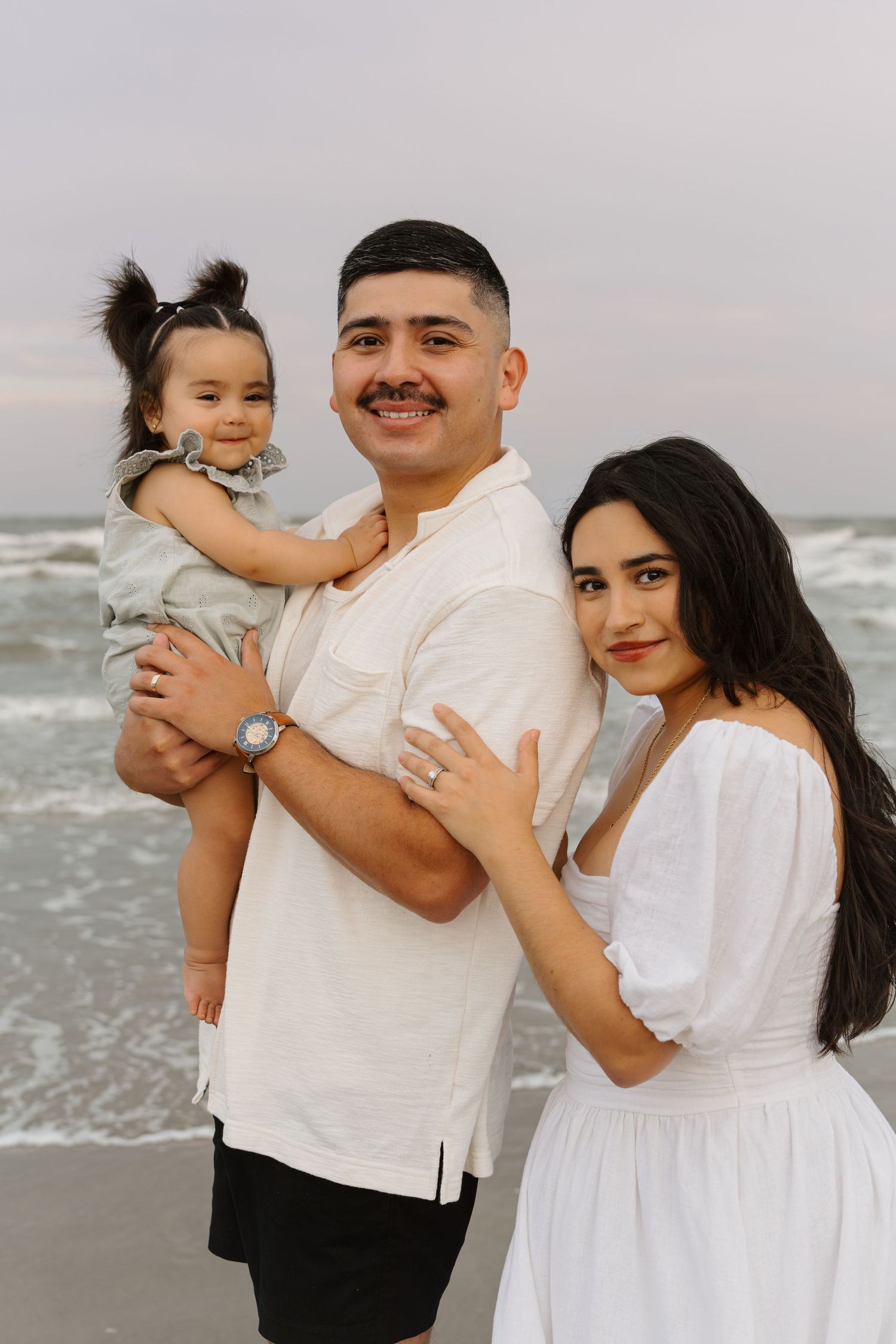 A young family of three poses on a beach, with a man holding a toddler beside a woman in a white dress.