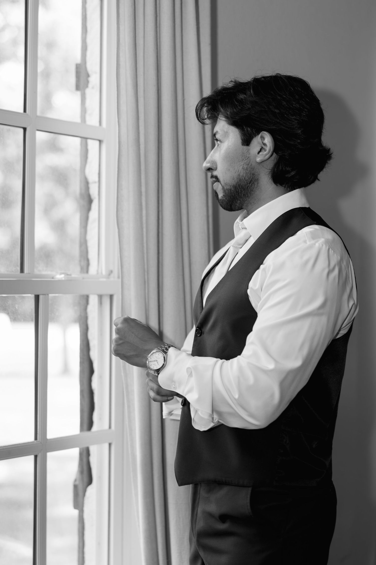 Black and white photo of groom in vest gazing thoughtfully out window before wedding ceremony.