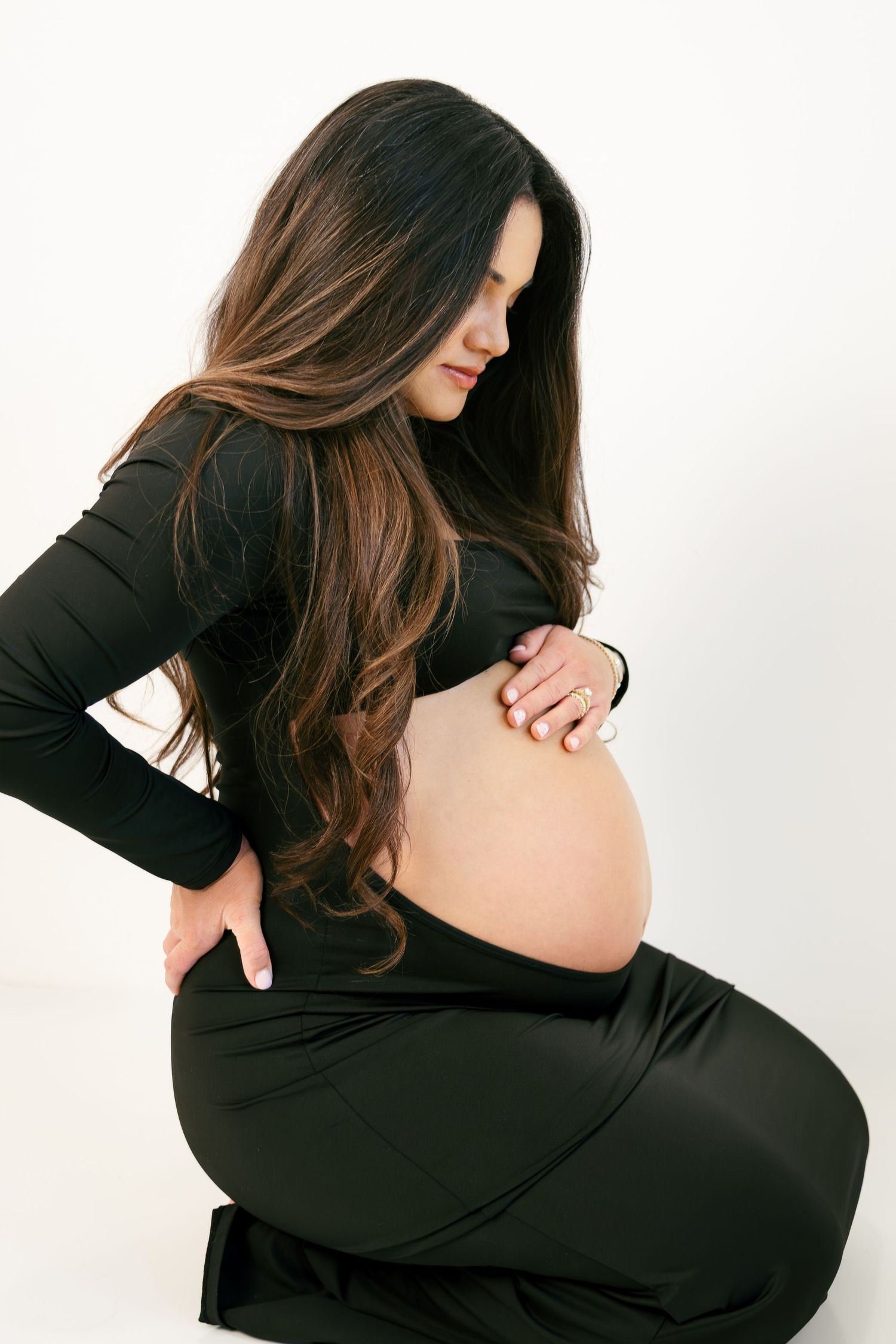 Pregnant woman in black outfit sitting and cradling her baby bump with long brunette hair against white background.
