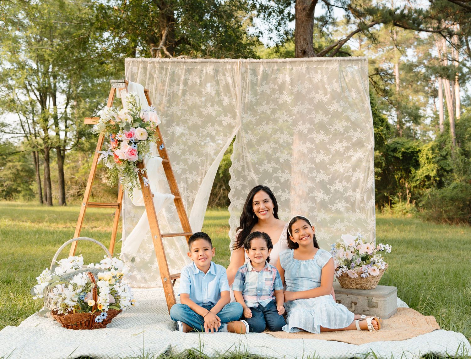 Family of four poses outdoors on a blanket before a lace backdrop with floral decor on a sunny day.