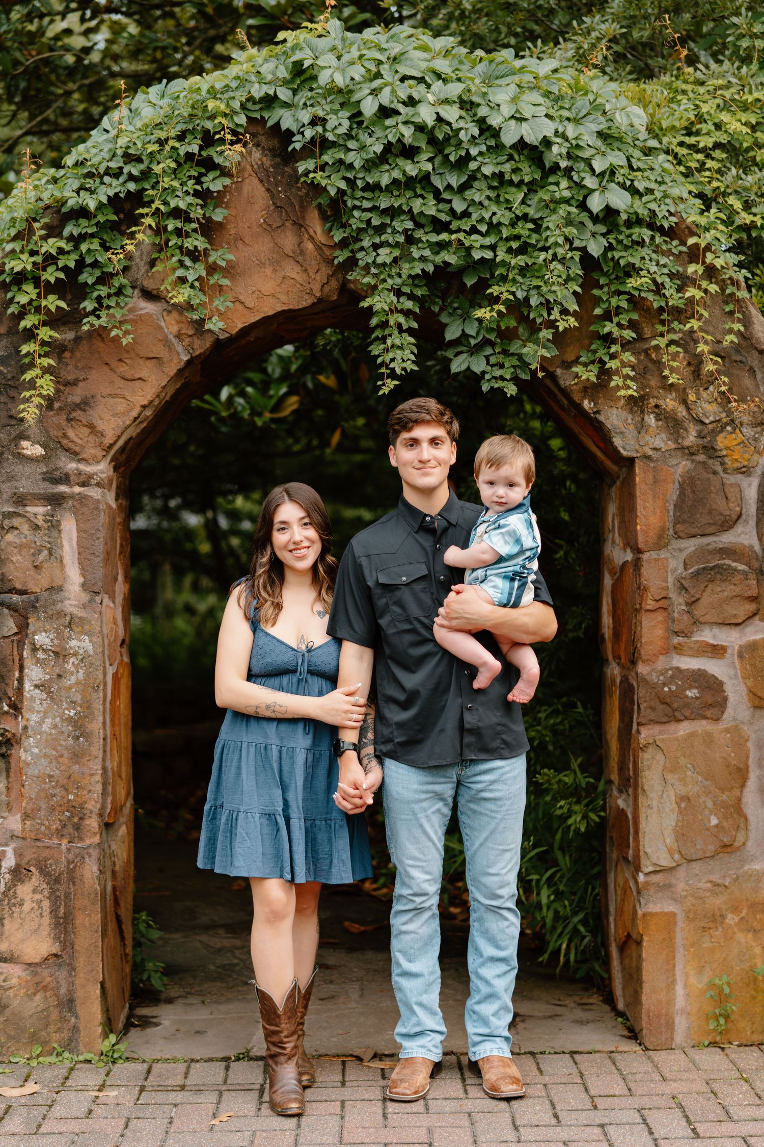 Young family of three posing under a stone arch covered in ivy, parents holding baby in a lush garden setting.