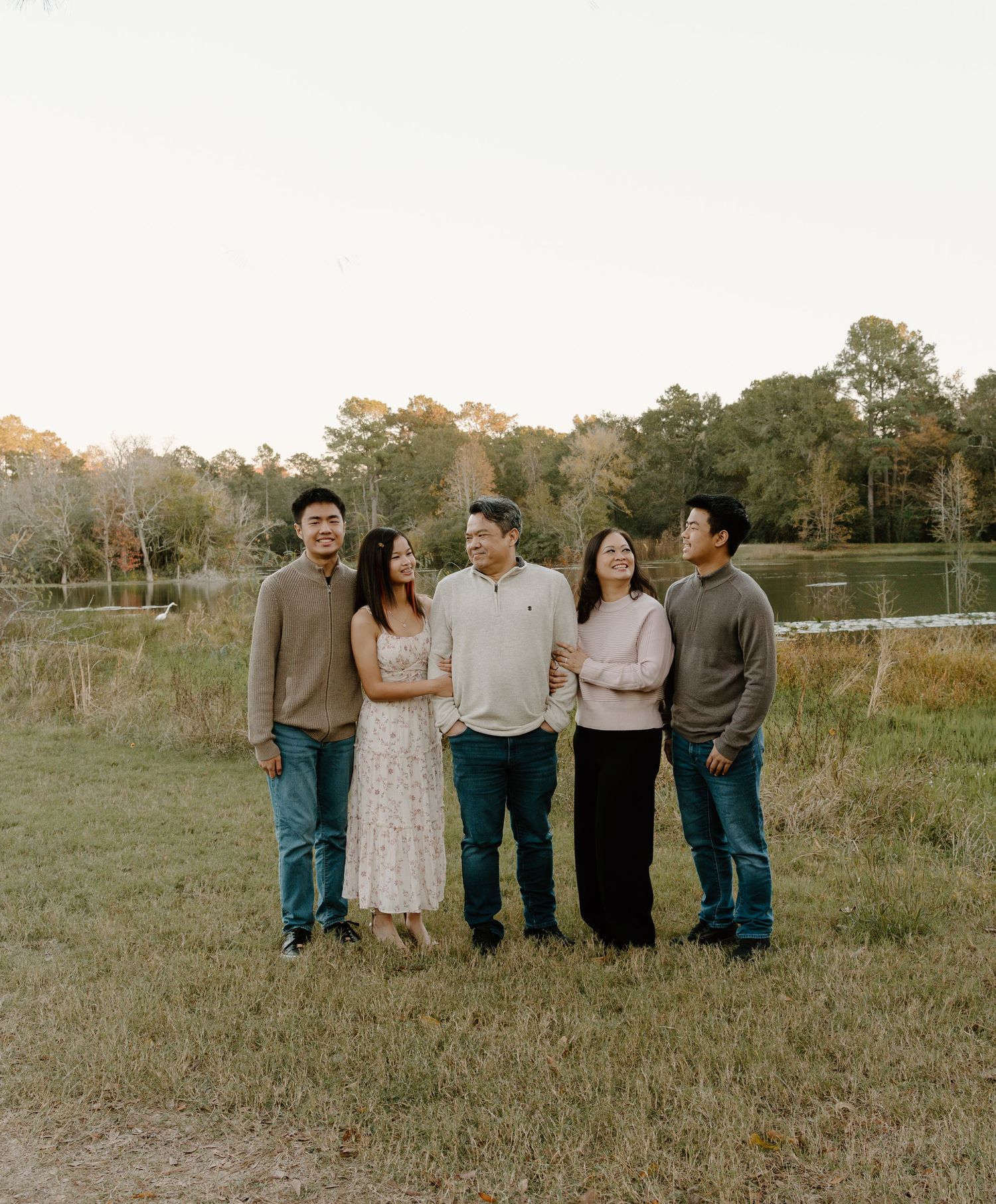 Five people smiling and posing together outdoors near a lake surrounded by trees during golden hour.