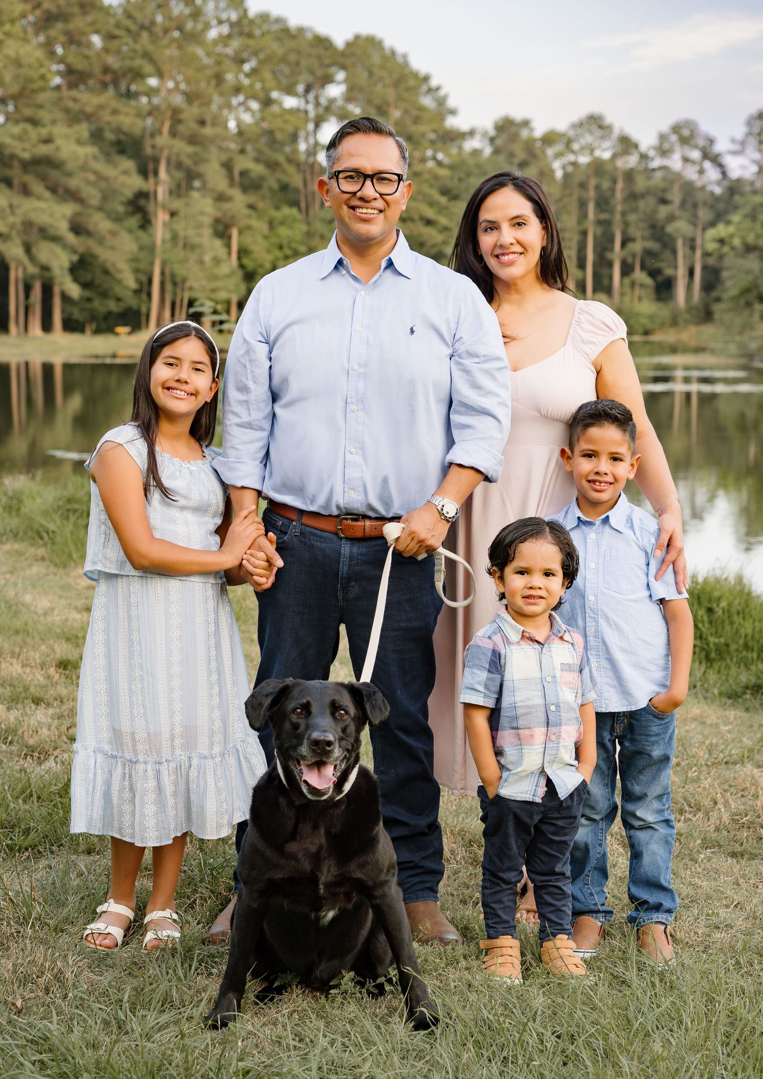Family of four poses outdoors with black Labrador near a pond surrounded by tall pine trees.
