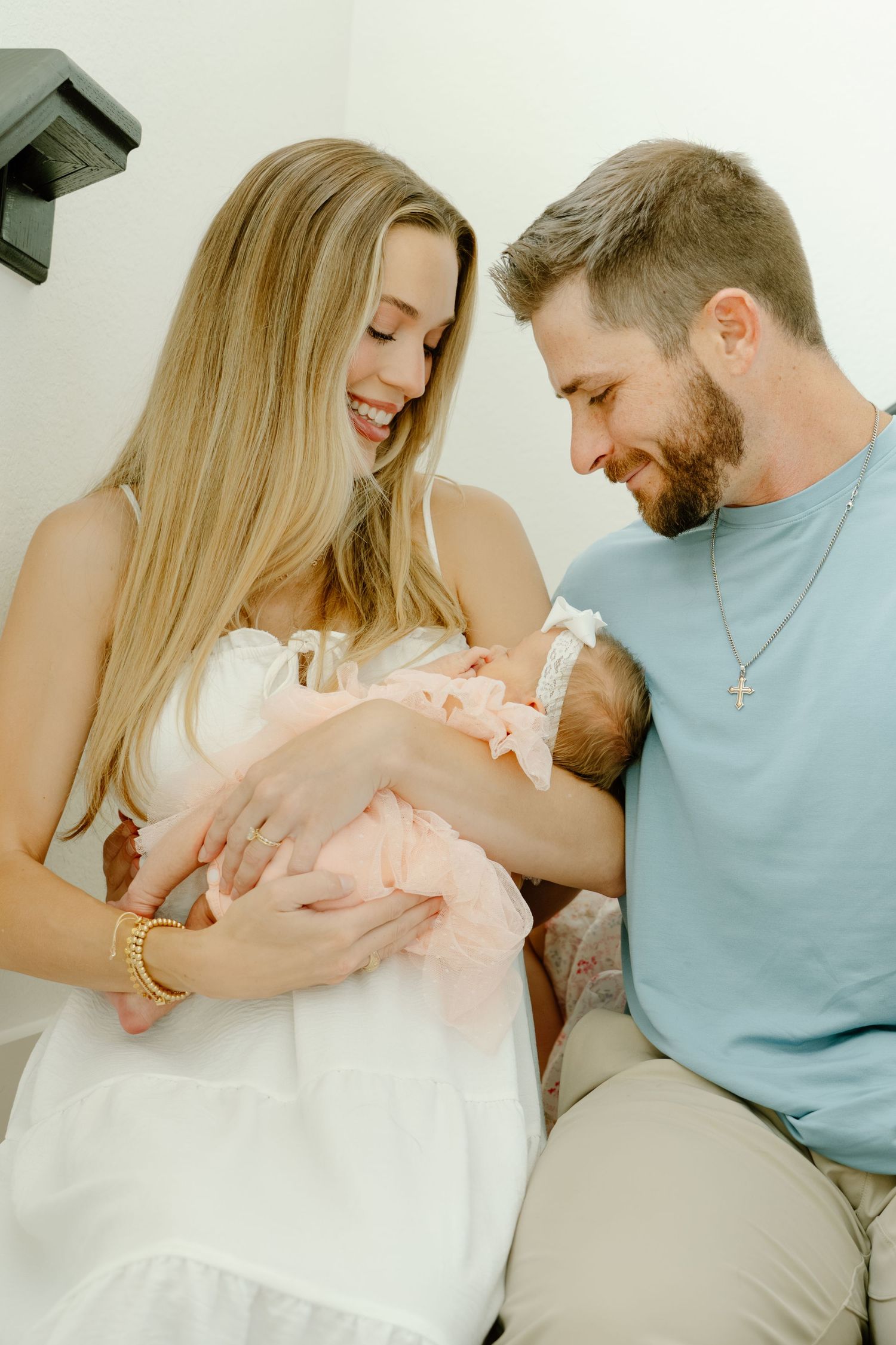 Happy couple smiling at their newborn baby, mother in white dress, father in blue shirt, bright indoor setting.