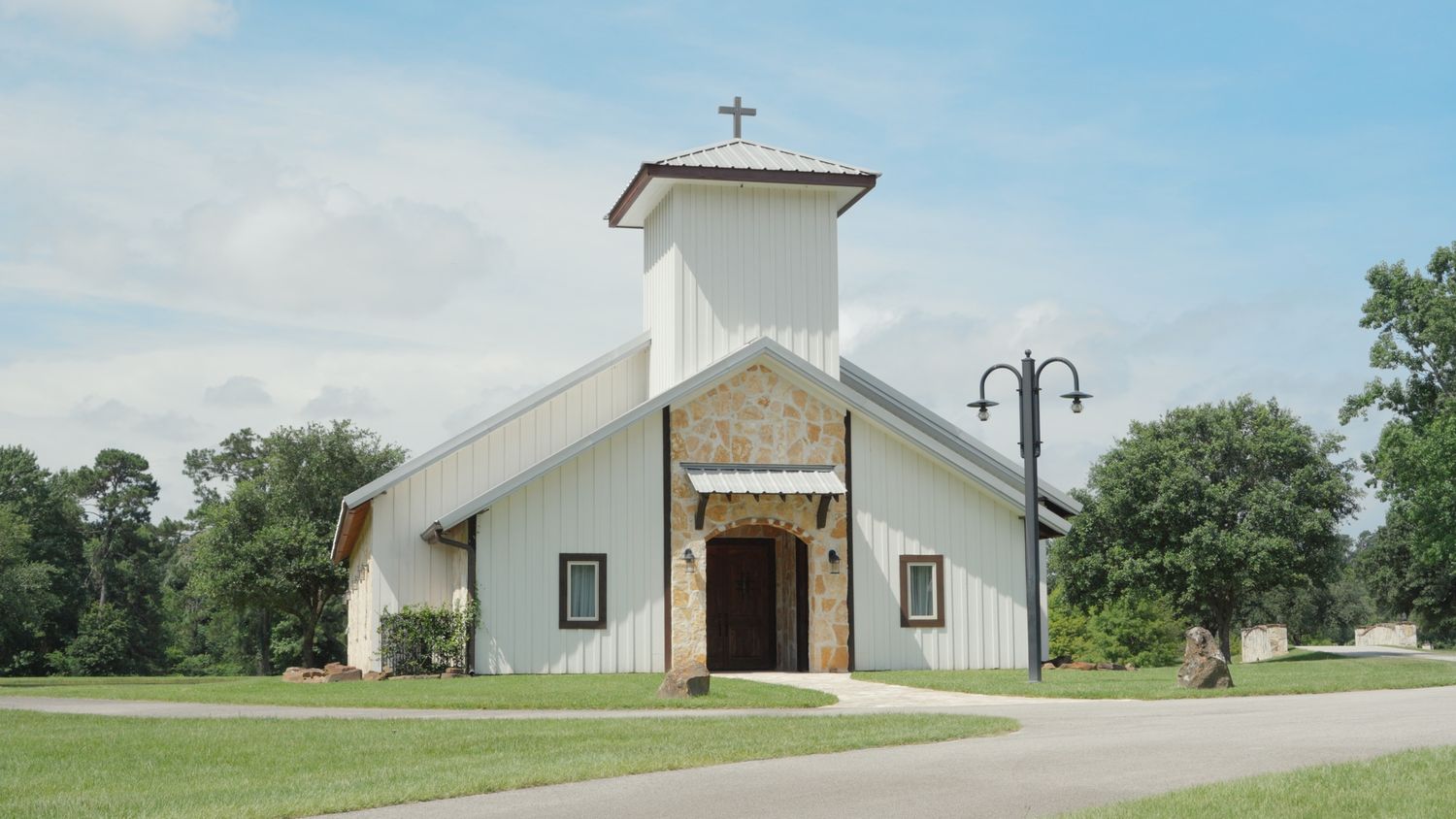 White country church with cross steeple surrounded by green trees and manicured lawn on a sunny day.