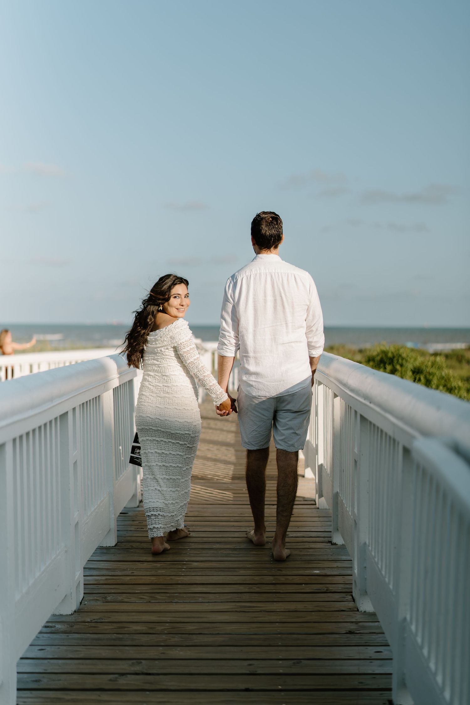 Couple in white outfits holding hands walking on a wooden boardwalk toward the beach on a sunny day.