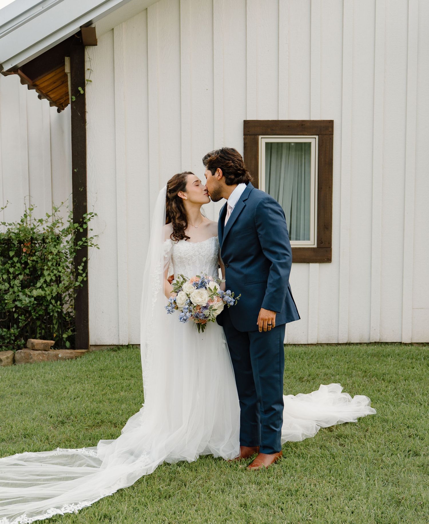 Newlyweds sharing a kiss outdoors on green lawn, bride in lace gown with long veil, groom in navy suit.
