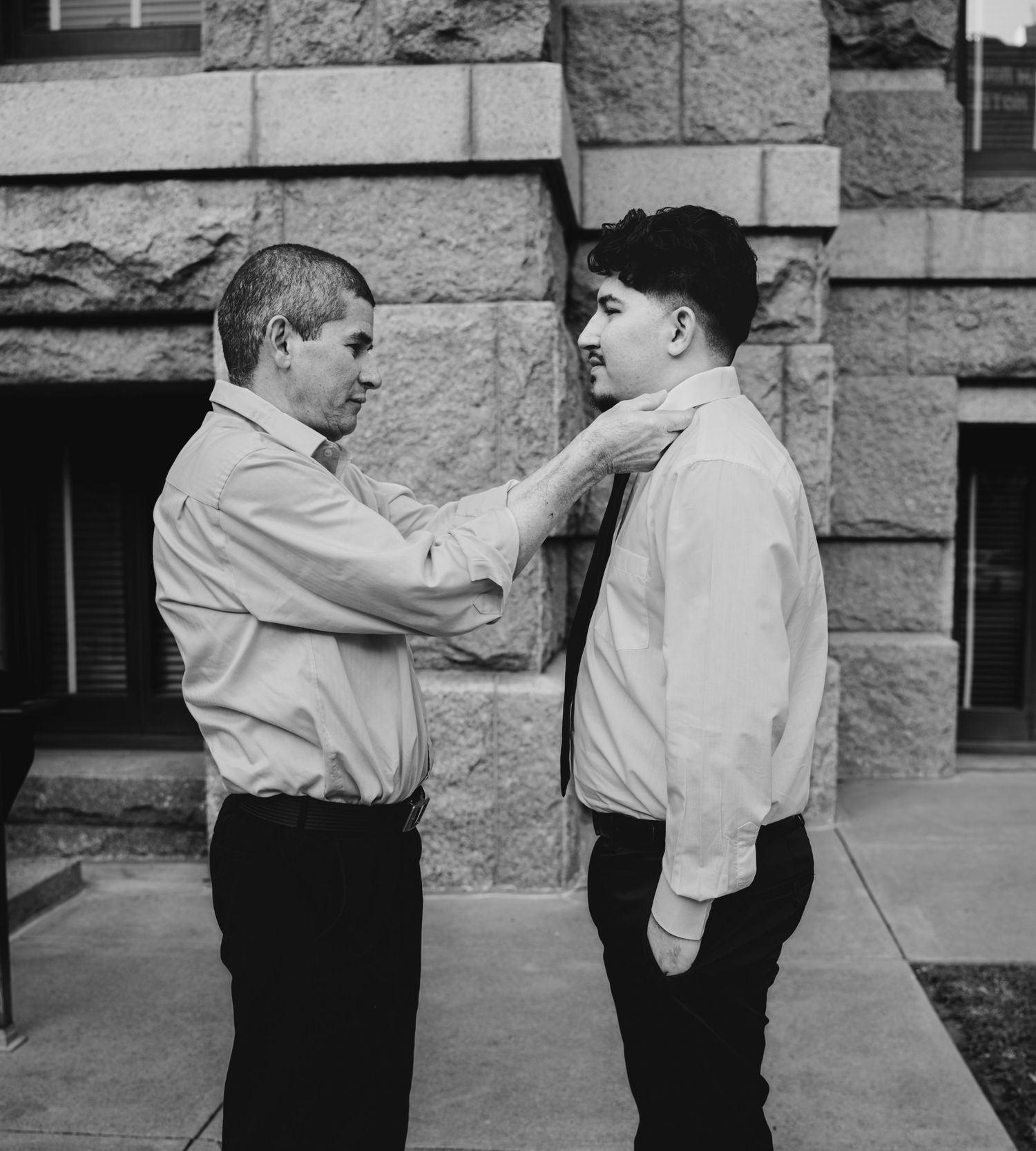 A black and white photo of a man adjusting another man's tie outside a stone building.