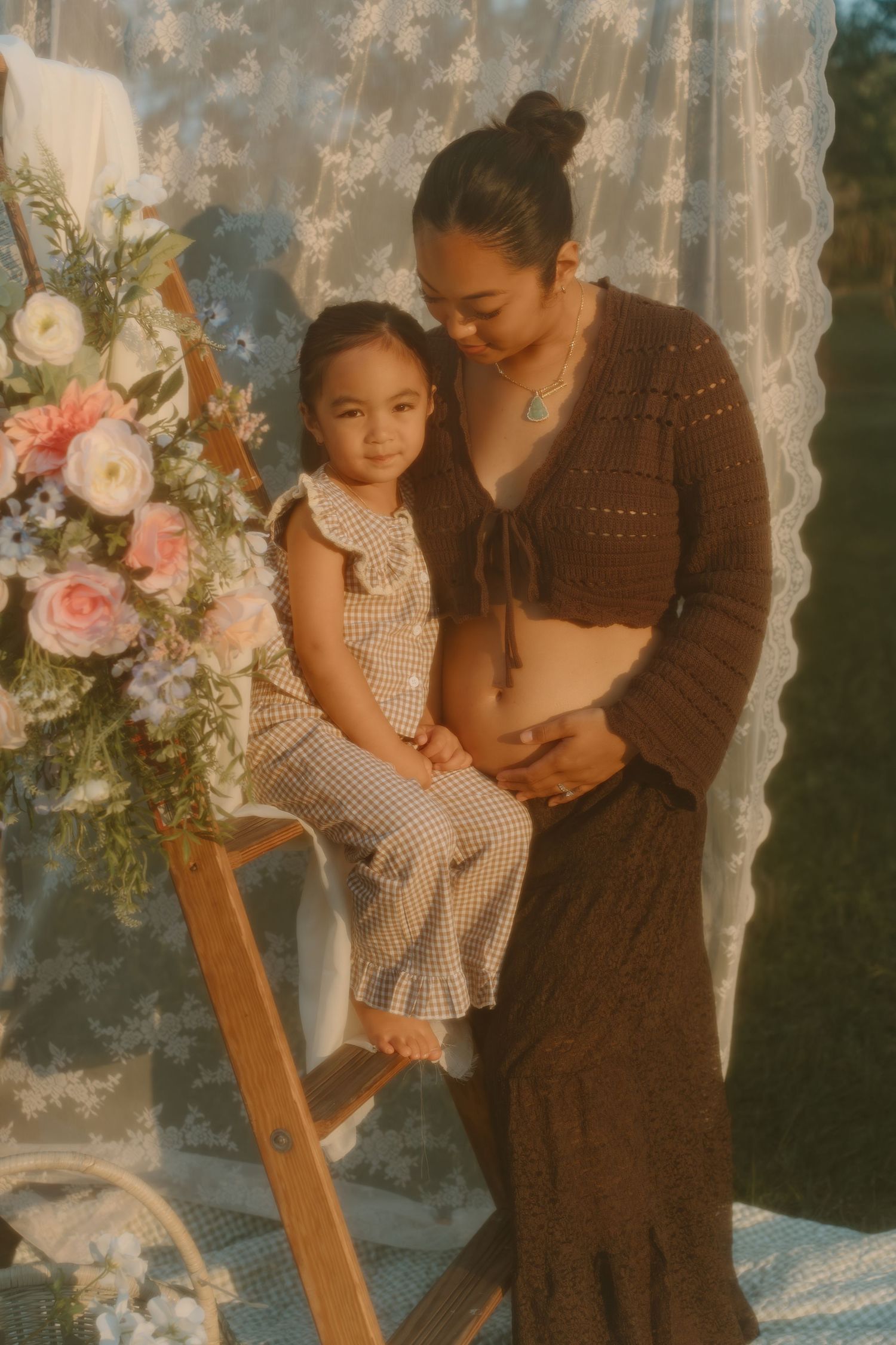 A pregnant woman in a brown crochet top embraces her young daughter on a ladder amid floral decor.
