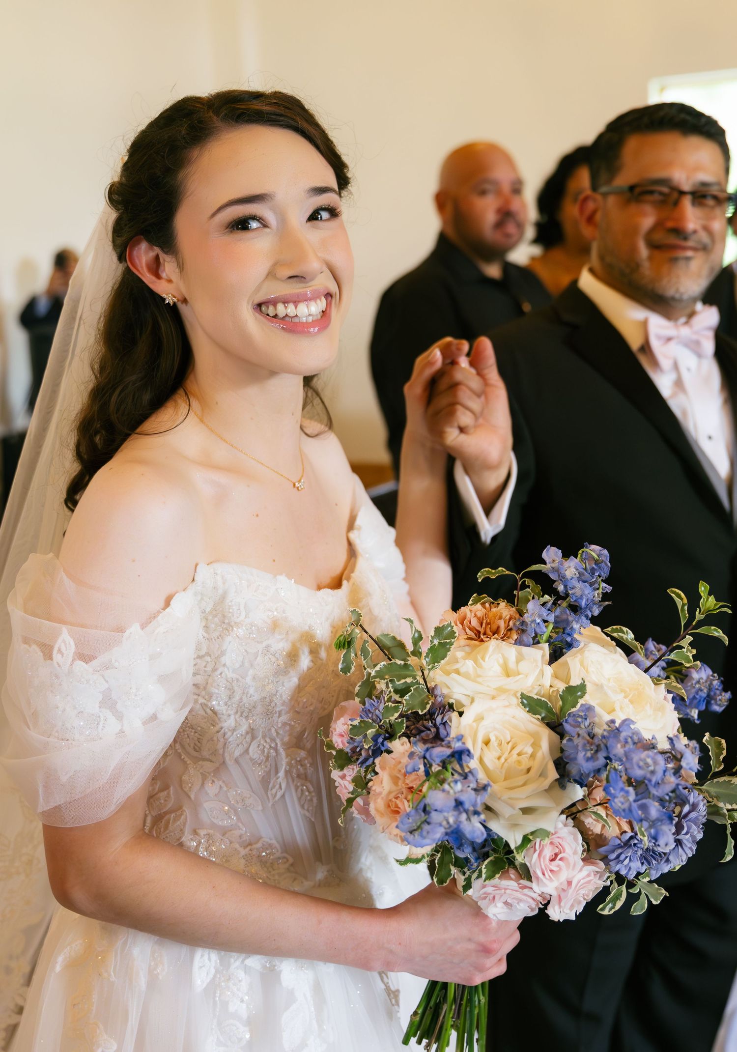 A radiant bride holding a blue and white floral bouquet smiles joyfully while walking down the aisle.