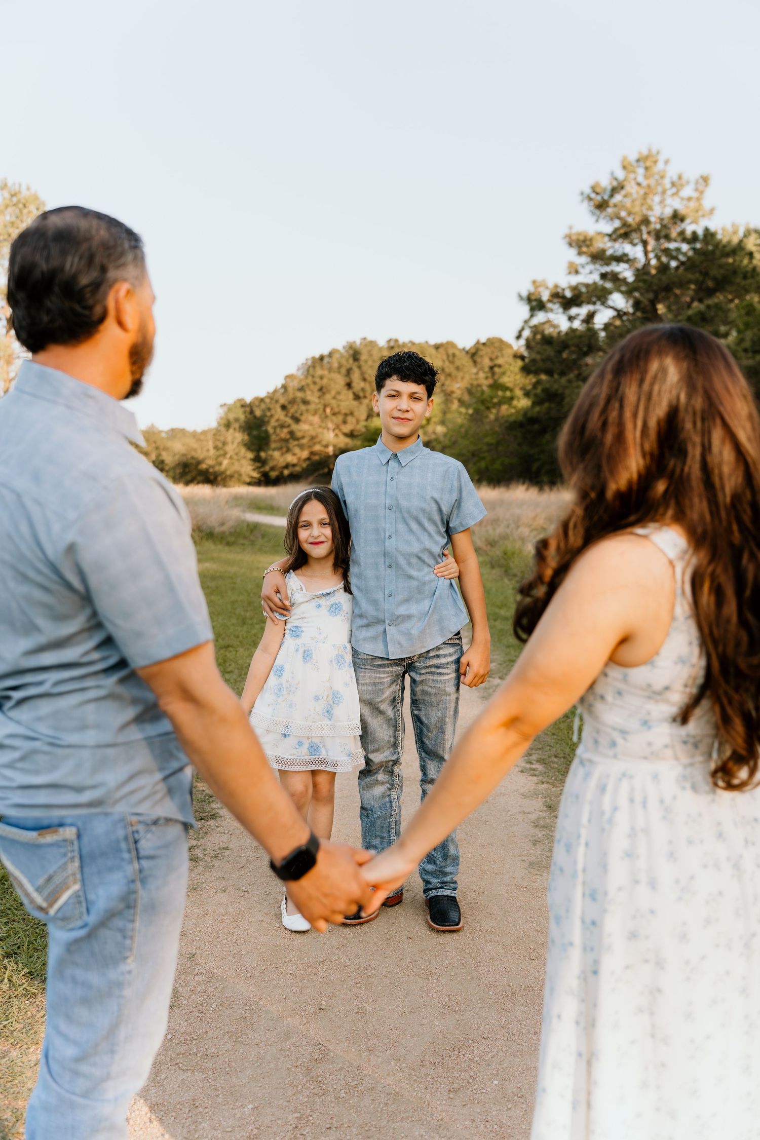 Family of four holding hands on a scenic dirt path at golden hour, with two children smiling at the camera.