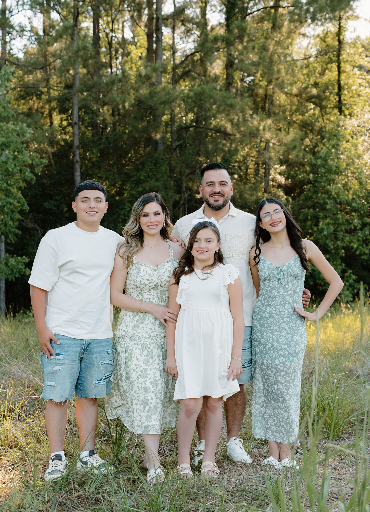 Family of five posing outdoors in coordinating green and white outfits surrounded by lush trees.