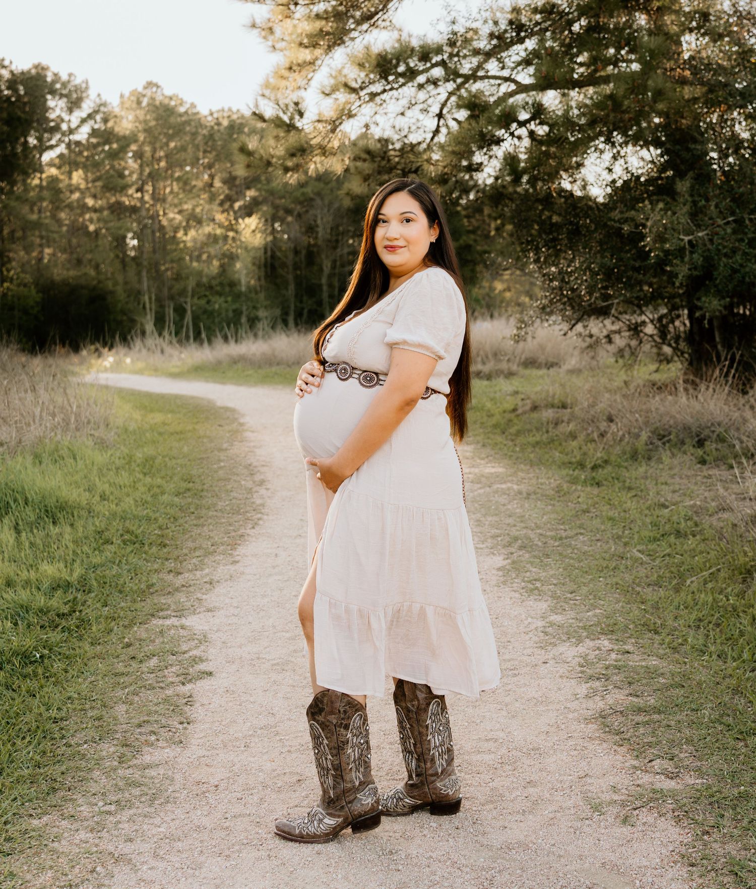 Pregnant woman in white dress and cowboy boots posing on scenic dirt path.