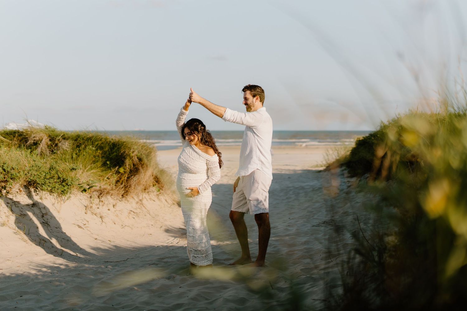 Couple dancing on a sandy beach path surrounded by dunes and ocean in matching white outfits.