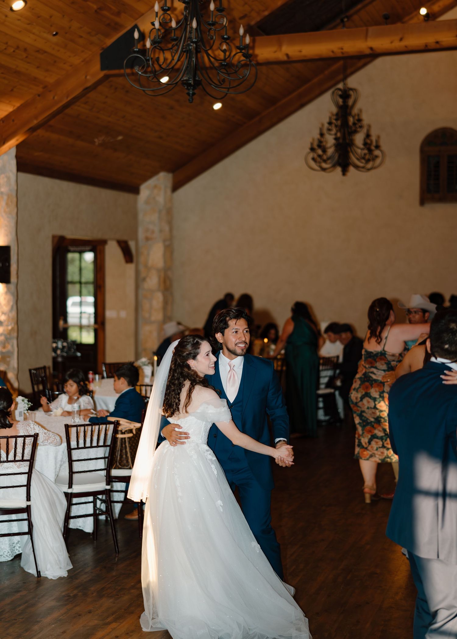 Bride and groom sharing first dance in rustic barn reception hall with chandeliers and seated guests.