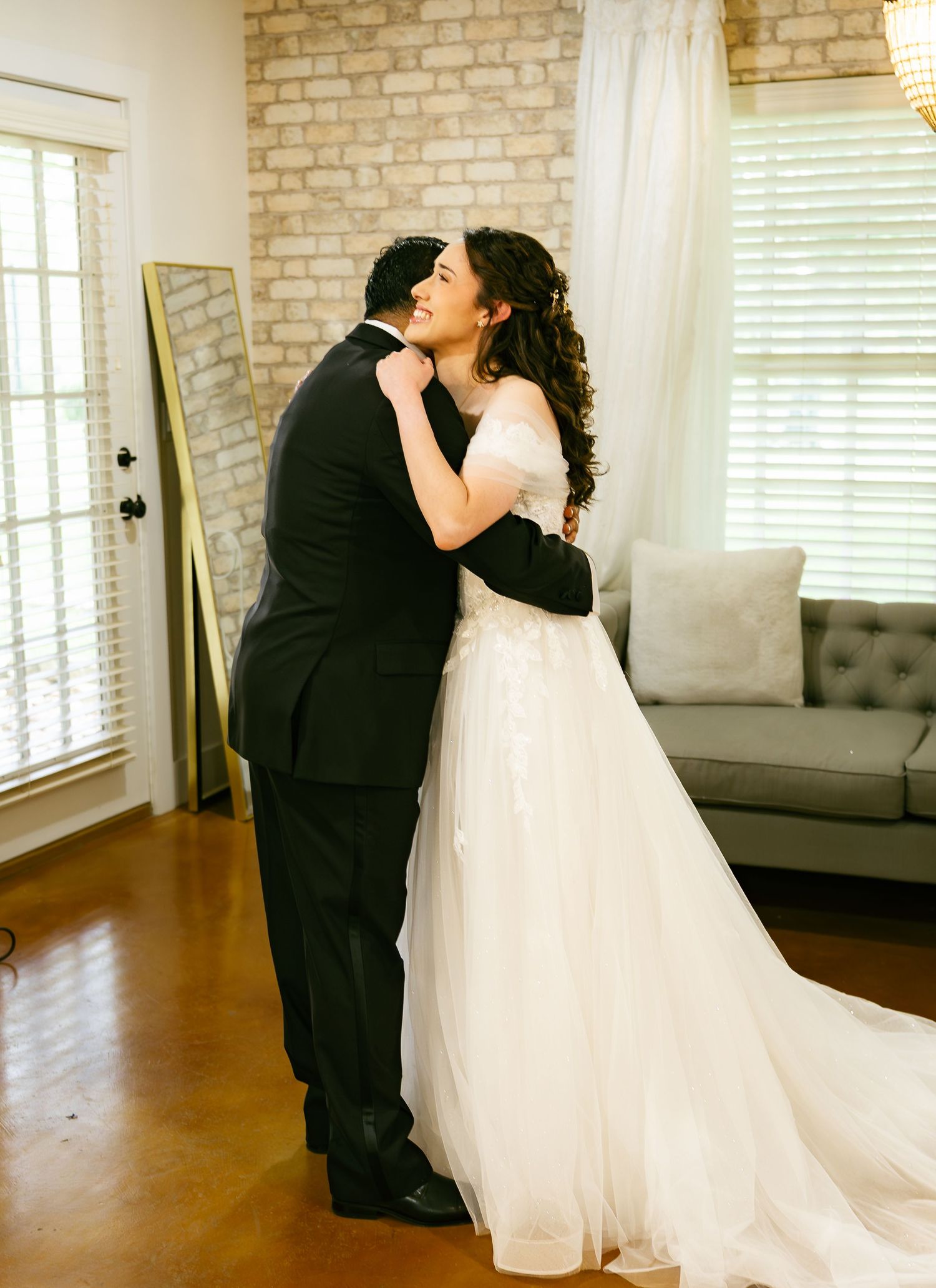 Emotional first look moment as groom in black tuxedo embraces bride in off-shoulder white wedding gown indoors.