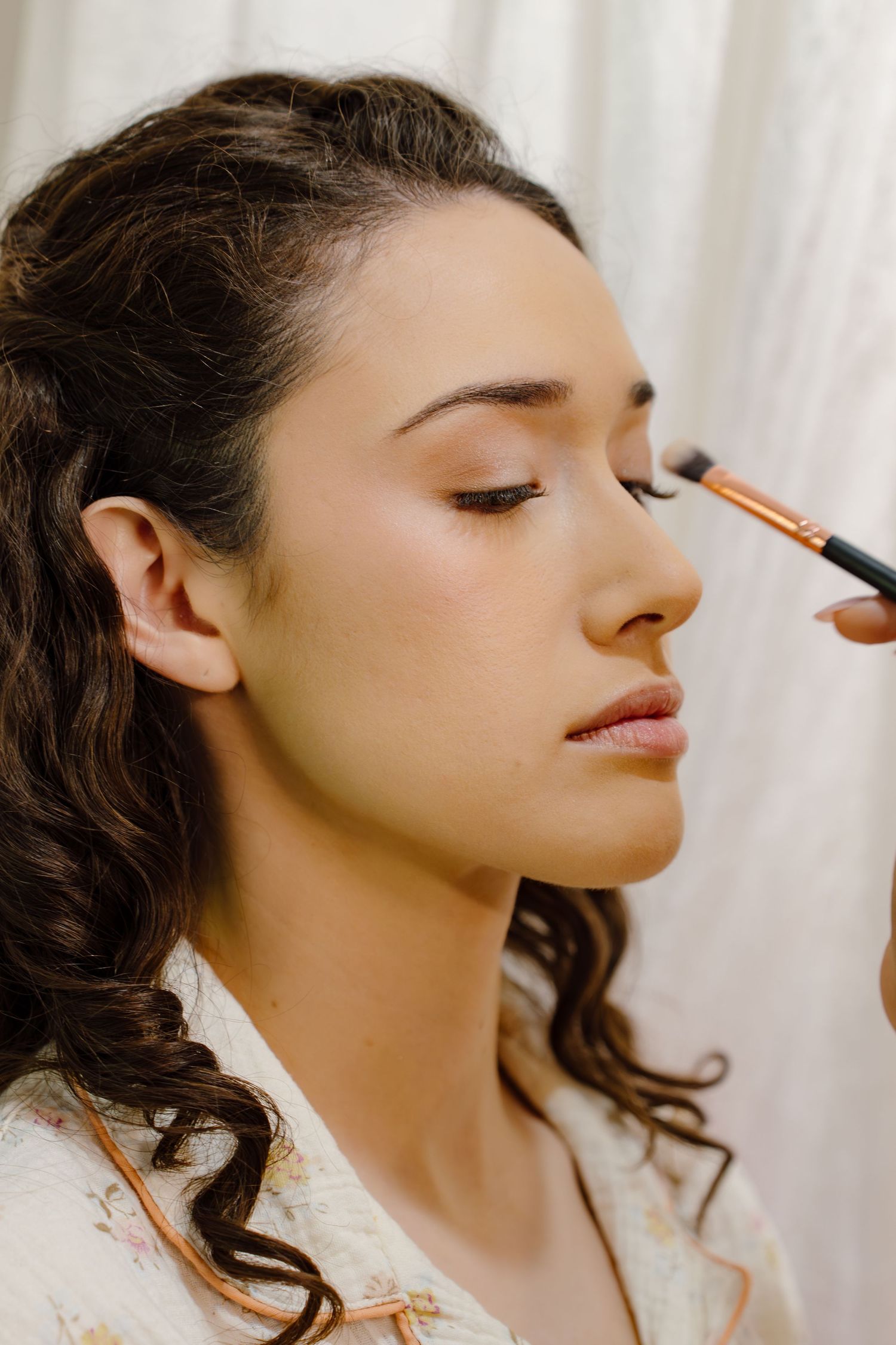 A makeup artist applies eyeshadow with a brush to a young brunette woman with curly hair in a floral robe.