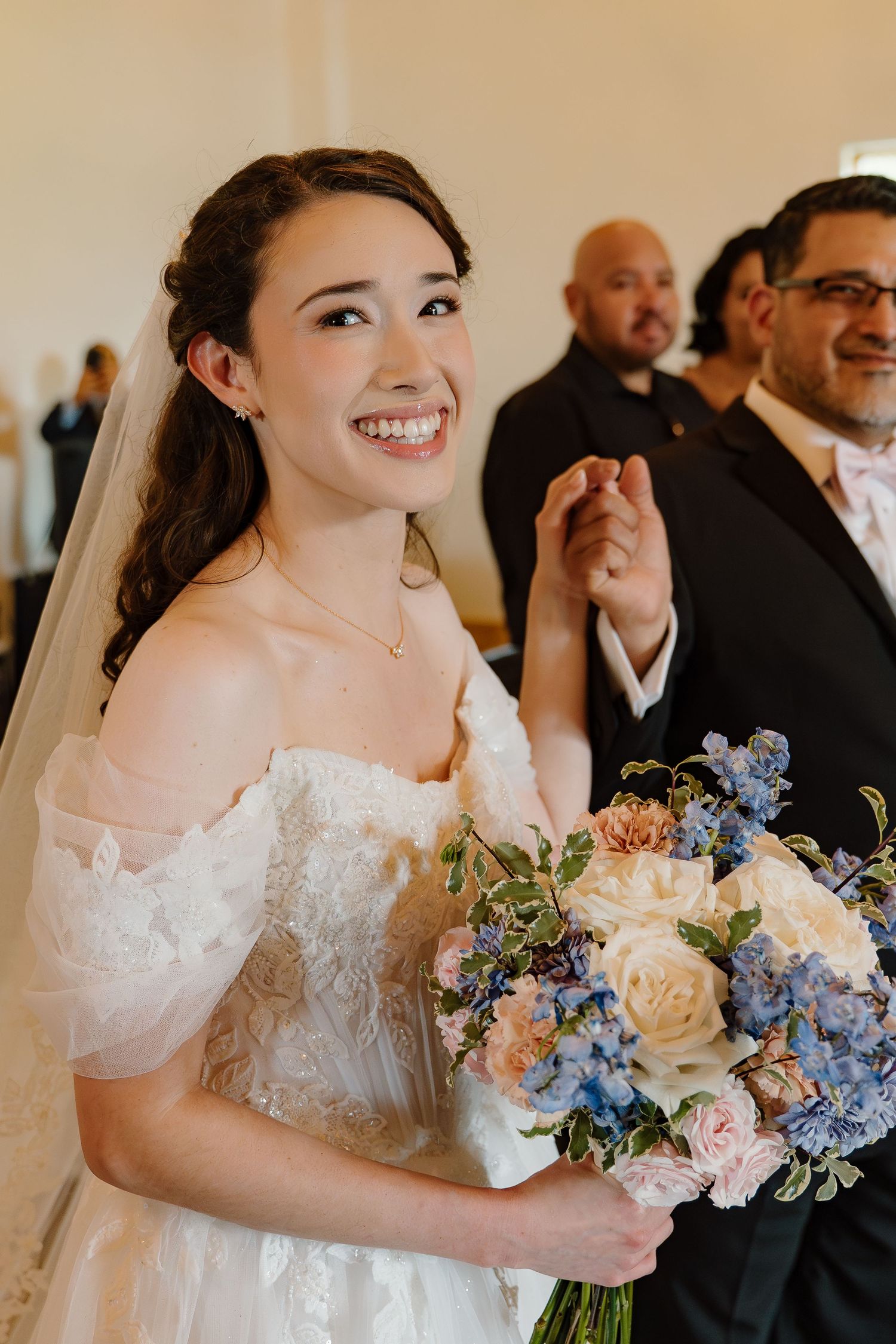 Smiling bride in off-shoulder lace gown holding blue and ivory bouquet walking down church aisle on wedding day.