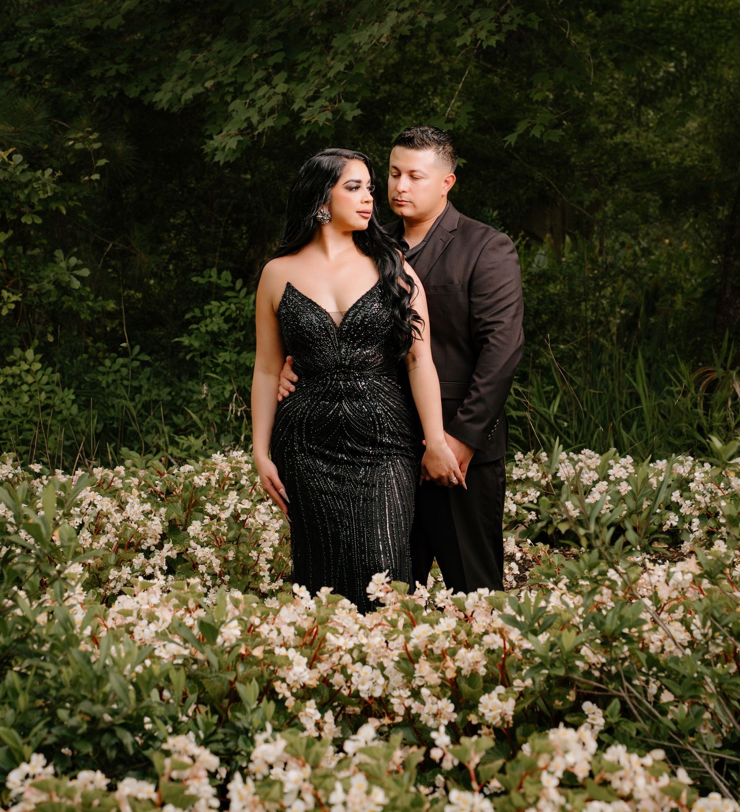 Couple in elegant black attire posing among white blooming flowers with lush green trees in background.