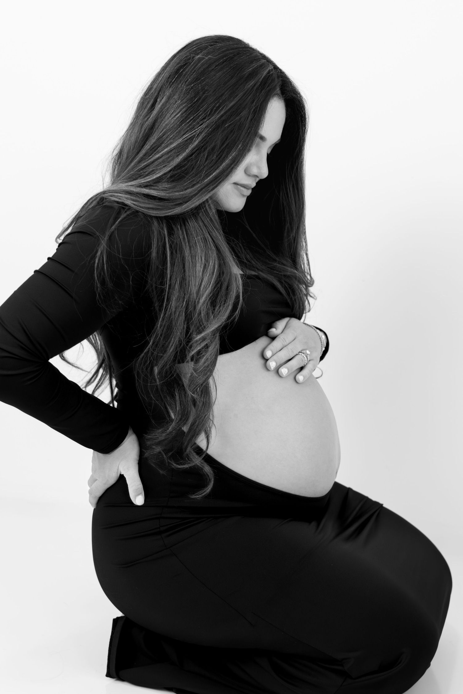 Black and white photo of a pregnant woman in black outfit cradling her bare baby bump while sitting.