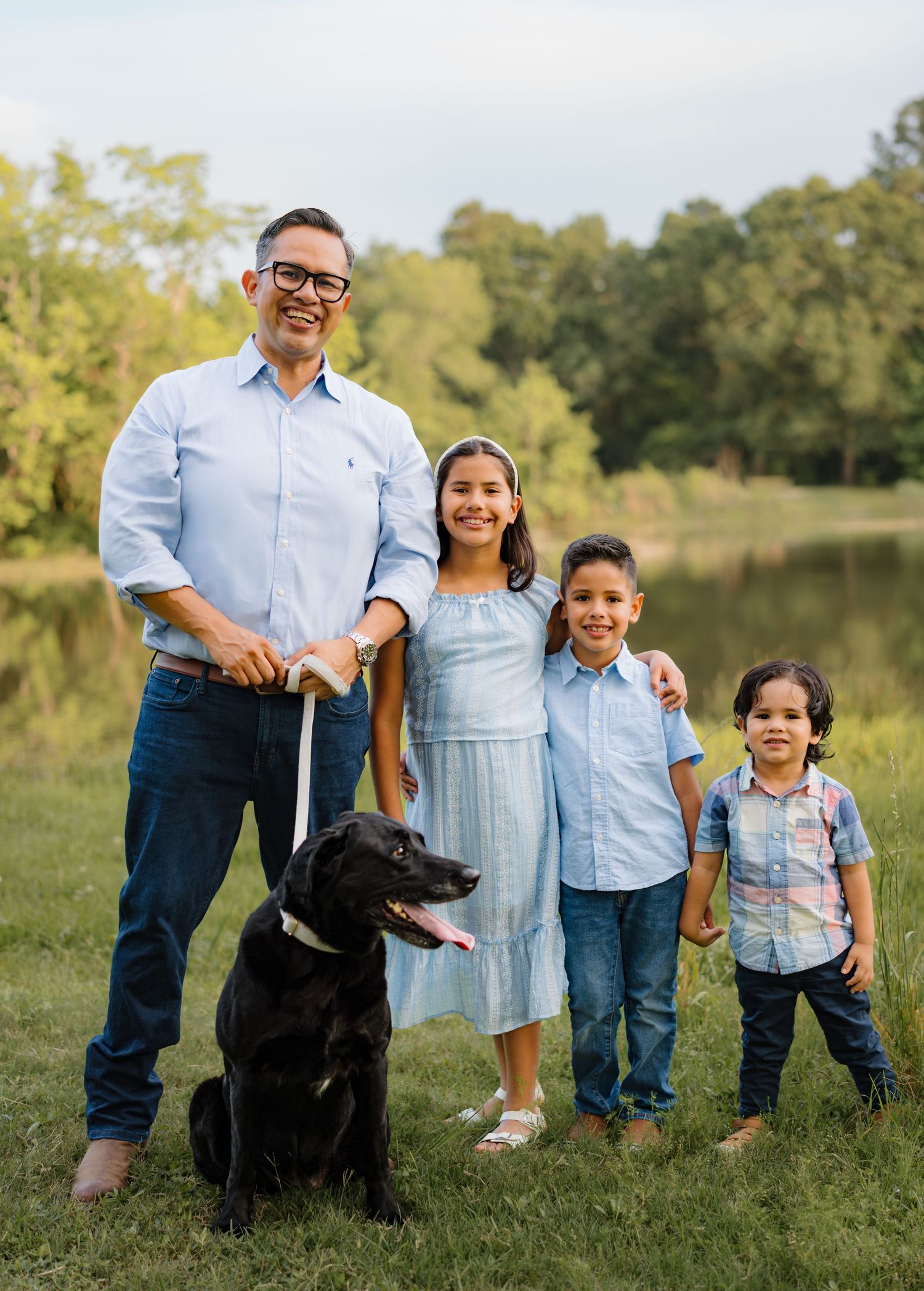 Family of four with a black Labrador dog posing outdoors near a pond surrounded by lush green trees.