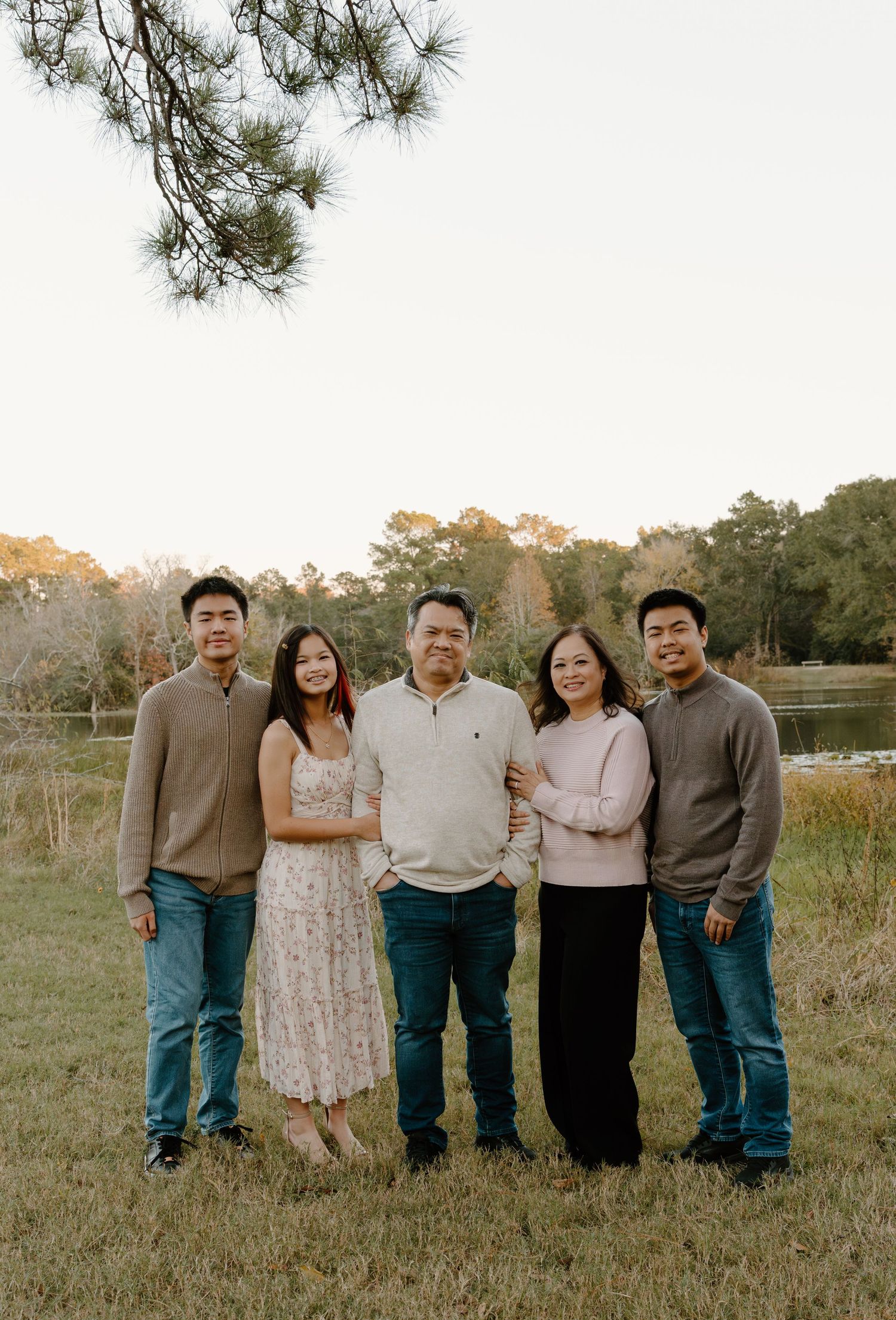 Family of five smiling outdoors near a lake surrounded by trees during golden hour in a park setting.