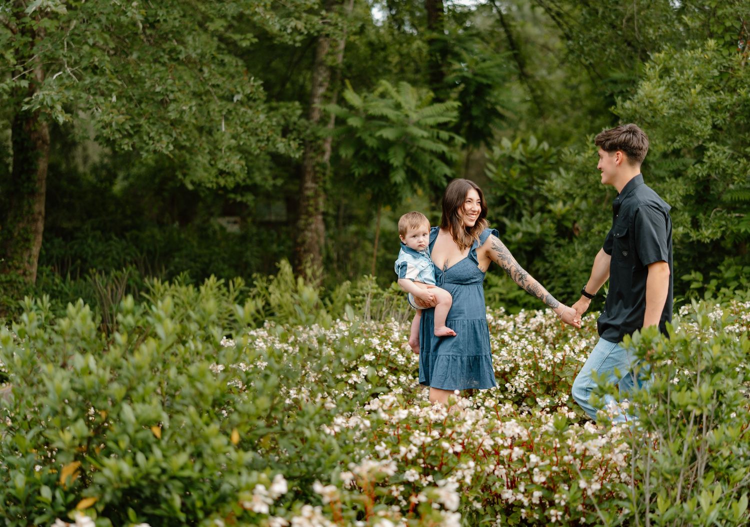 A young family walks through a lush garden, mother holding baby while father holds her hand among white flowers.