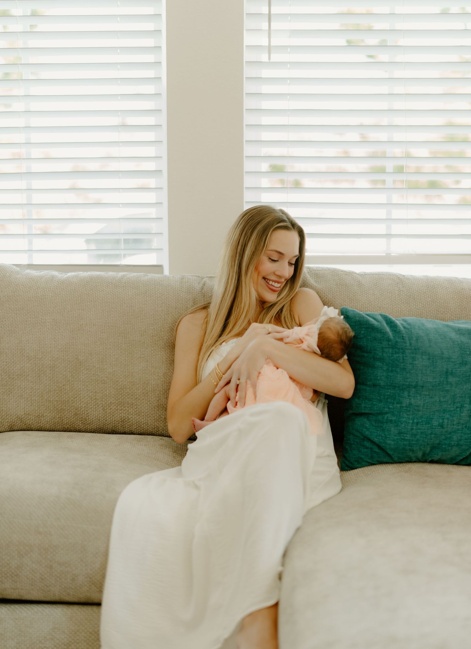 A smiling blonde mother sits on a beige sofa breastfeeding her newborn baby, wrapped in a white blanket.