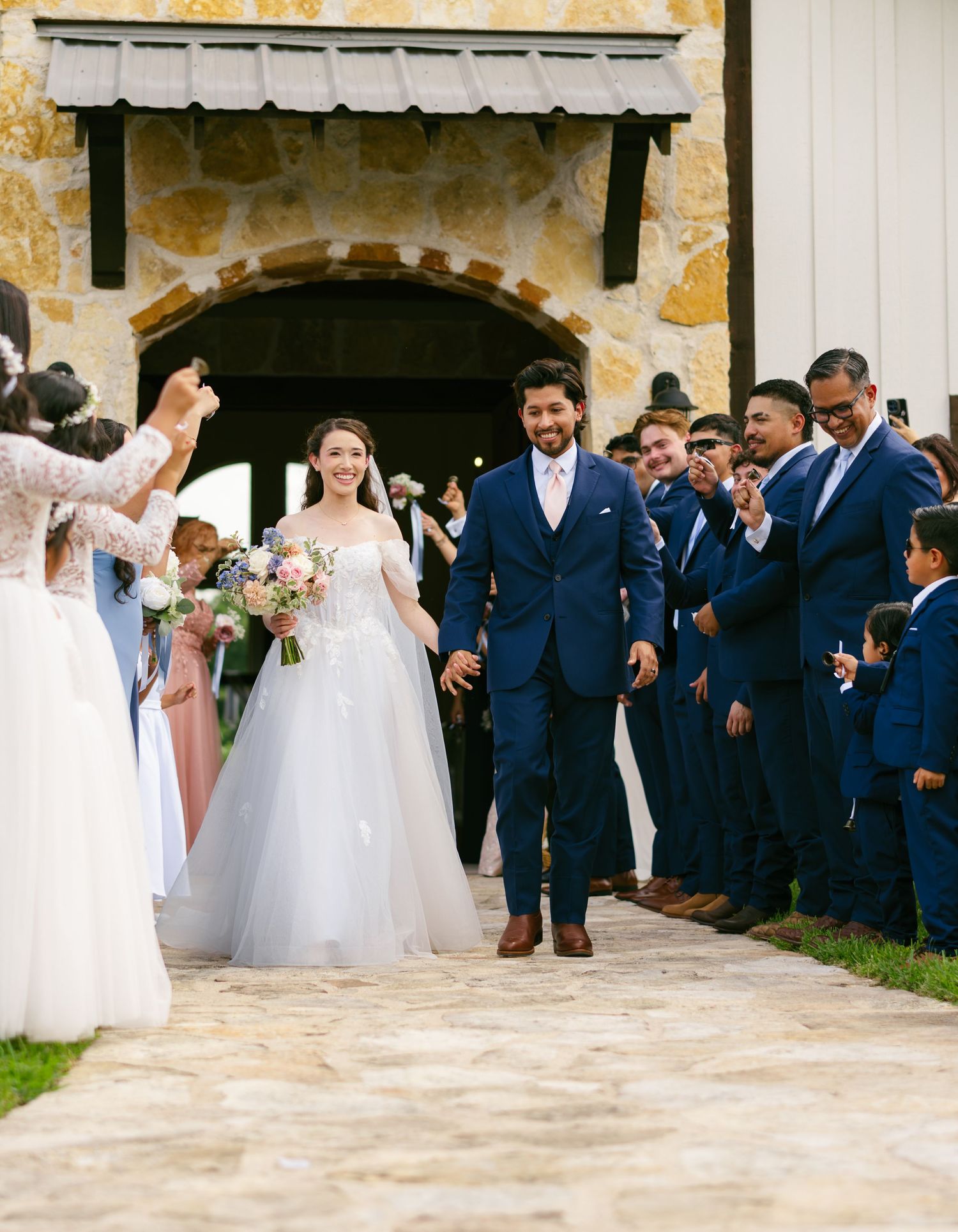 Bride and groom exit stone chapel, surrounded by wedding party in blue suits, as bride holds colorful bouquet.