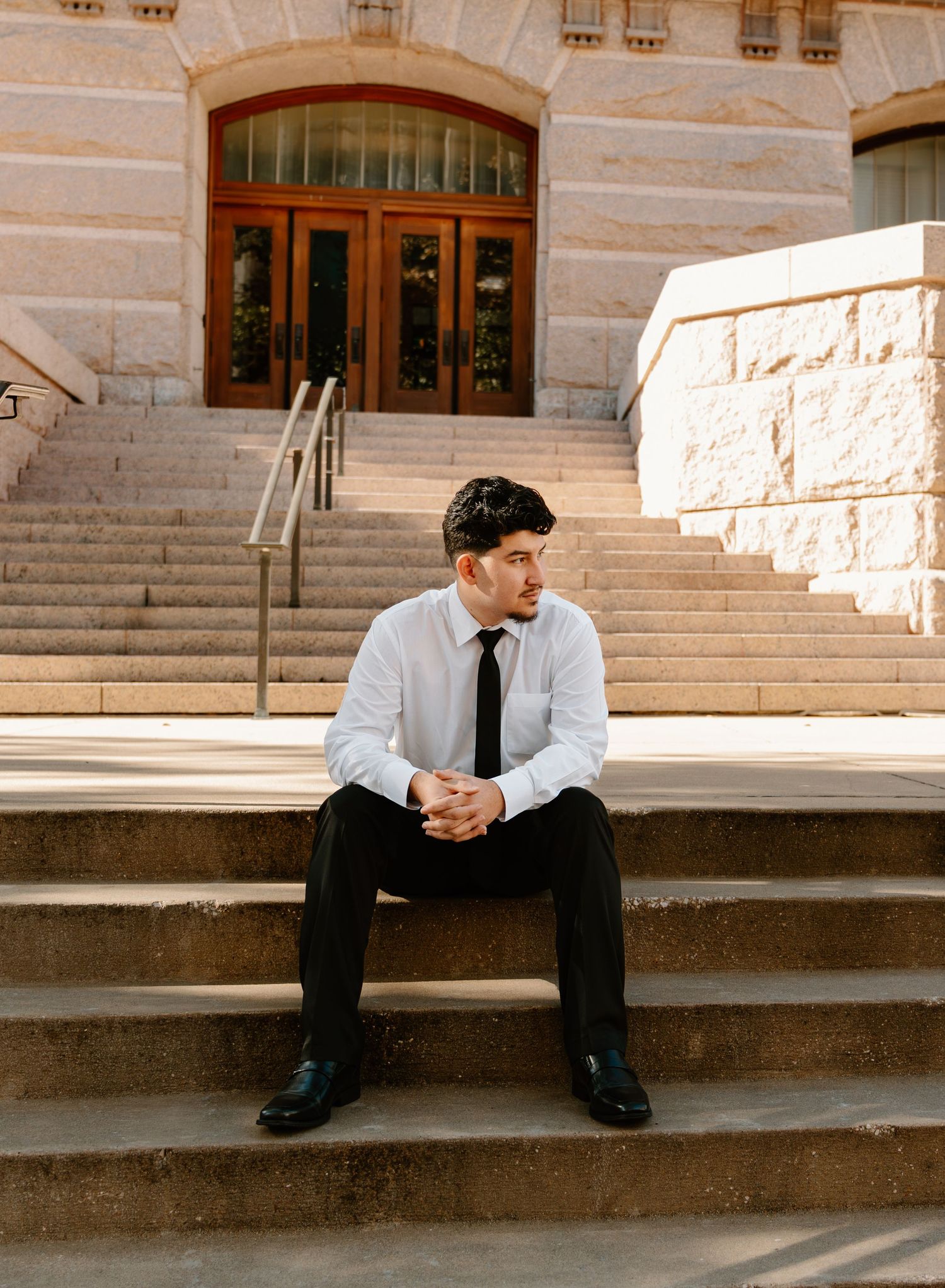 A well-dressed man in a white shirt and black tie sits thoughtfully on courthouse steps outdoors.