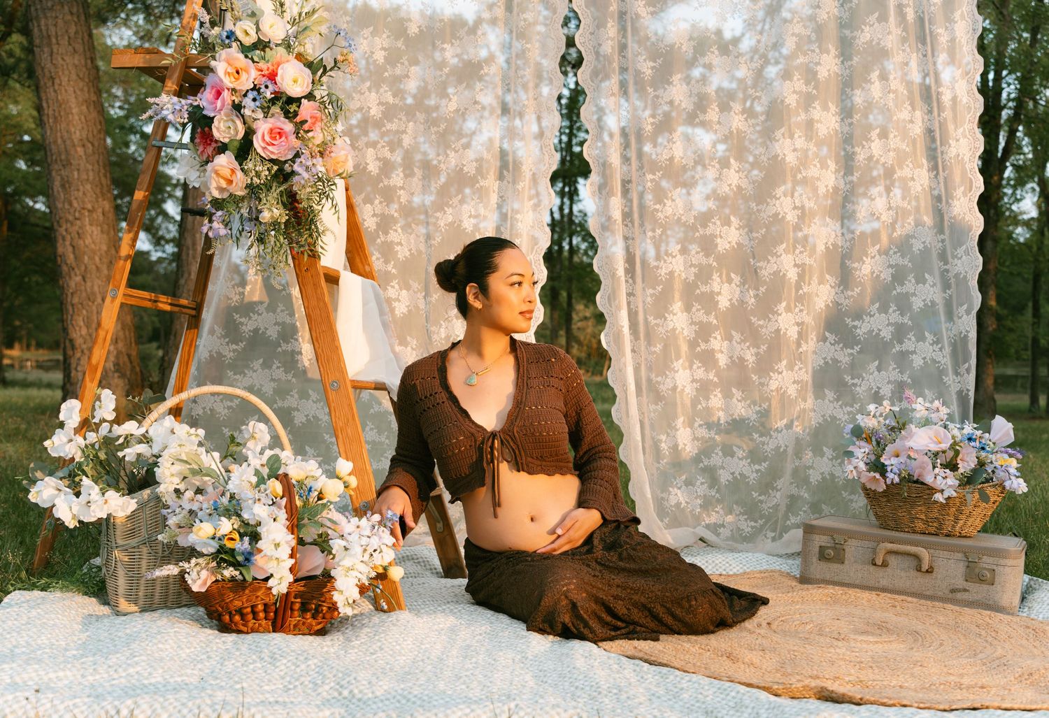 A pregnant woman in a brown crop top and skirt sits gracefully among flowers and lace decor outdoors.