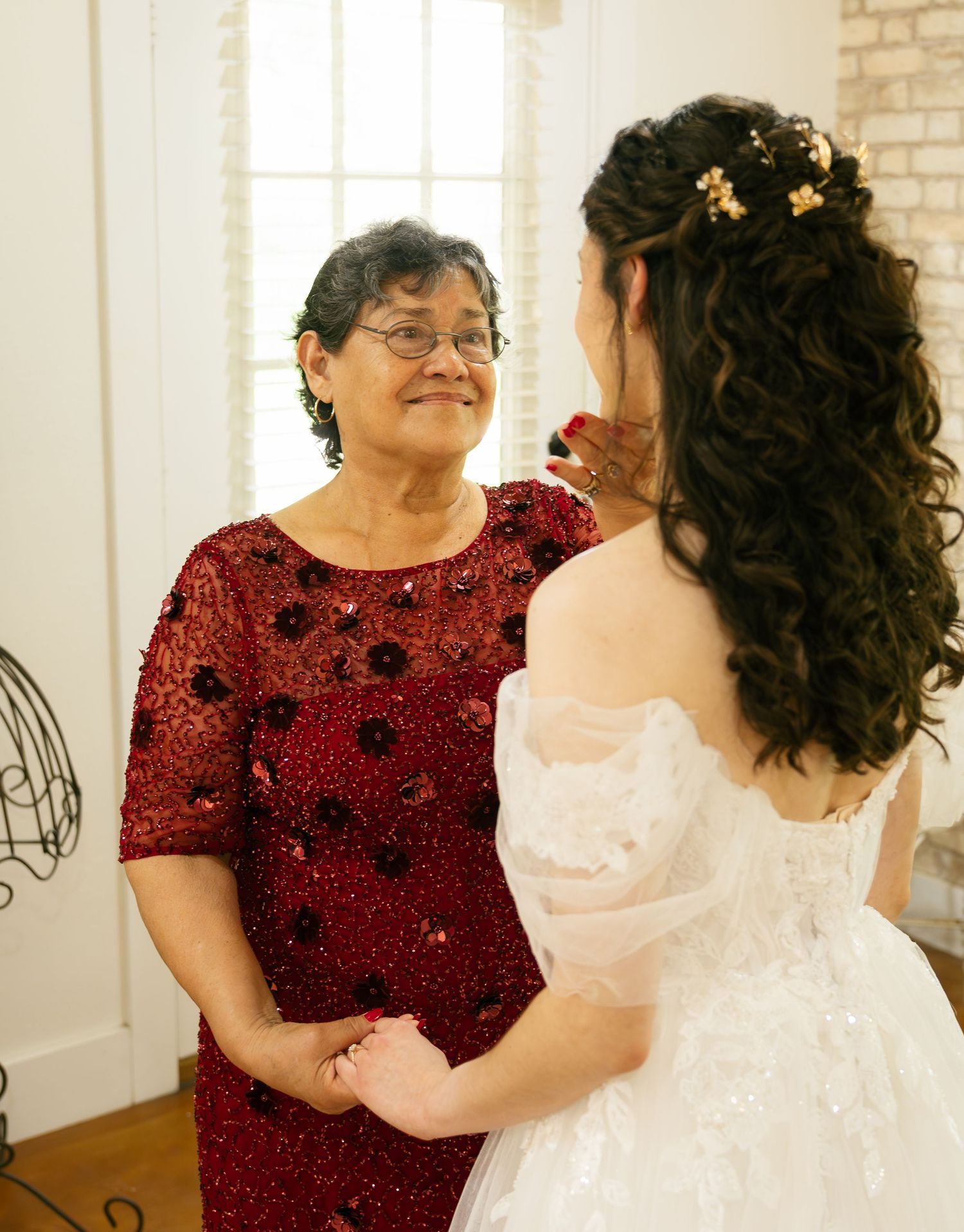 A bride in a white gown holds hands with an older woman in a red beaded dress, sharing a tender moment.